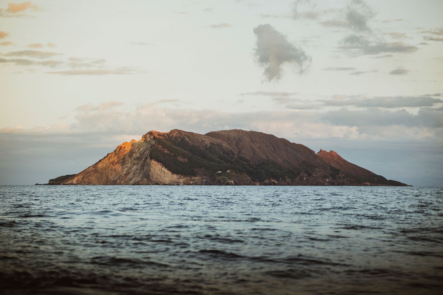 Rocky island rising from calm ocean waters at golden hour, with dramatic clouds in the pastel sky above.