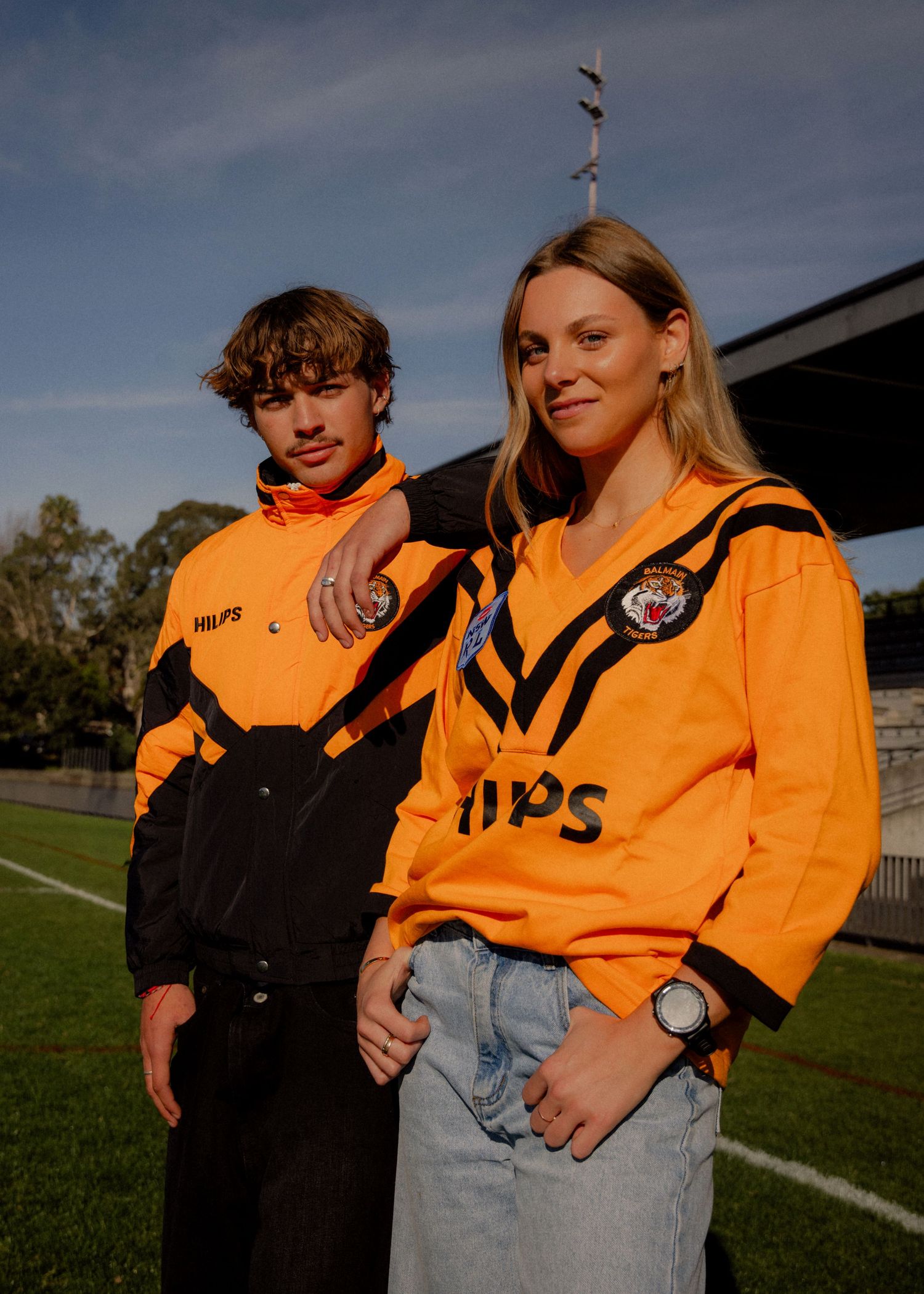 Two models wearing orange and black Wests Tigers NRL jerseys with Philips sponsor logo at a football stadium. Business and brand