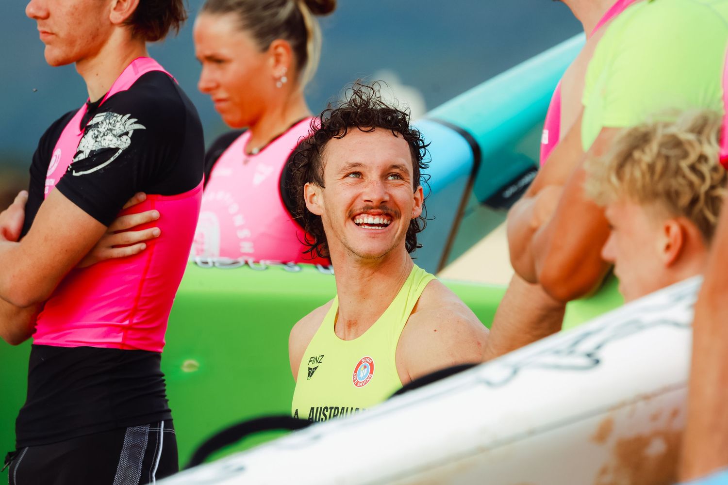 Smiling athlete in yellow Australia singlet stands among competitors holding colorful surfboards at a beach sports event.