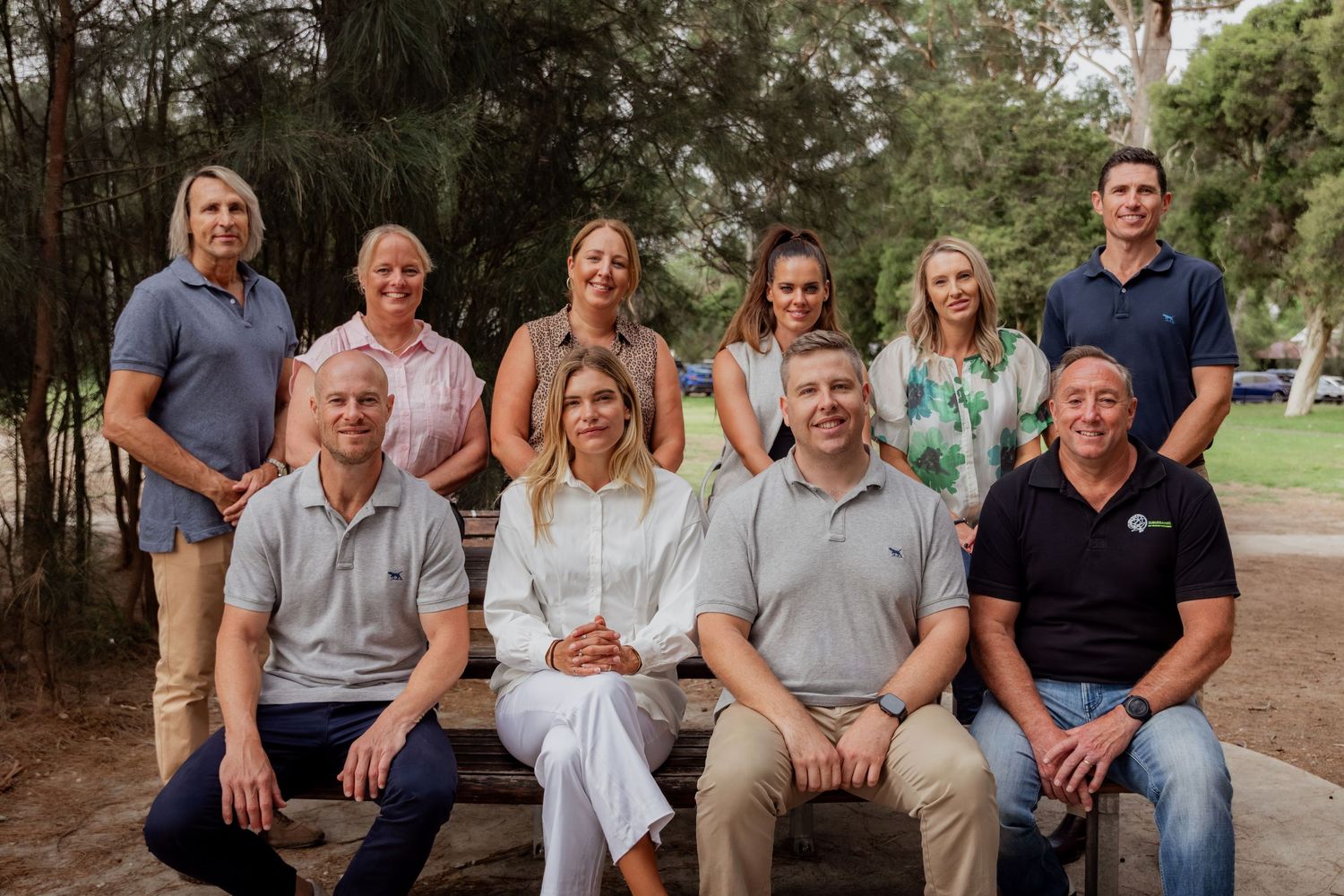 Group of ten professionals in polo shirts posing outdoors in a park setting for a team photo.