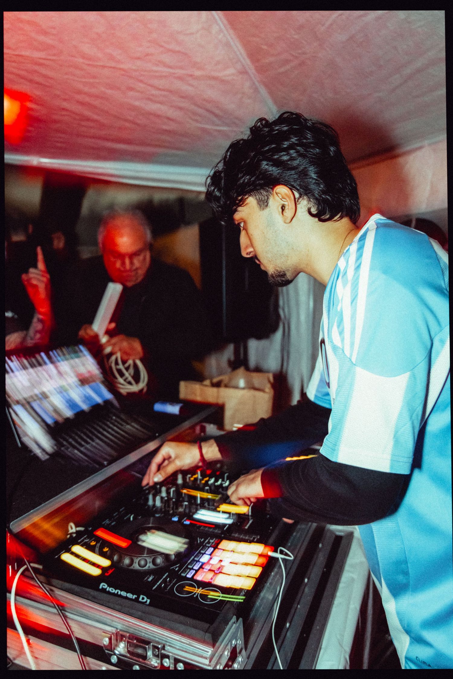 Young DJ mixing tracks on Pioneer DJ controller at a dimly lit event, wearing an Adidas jersey.