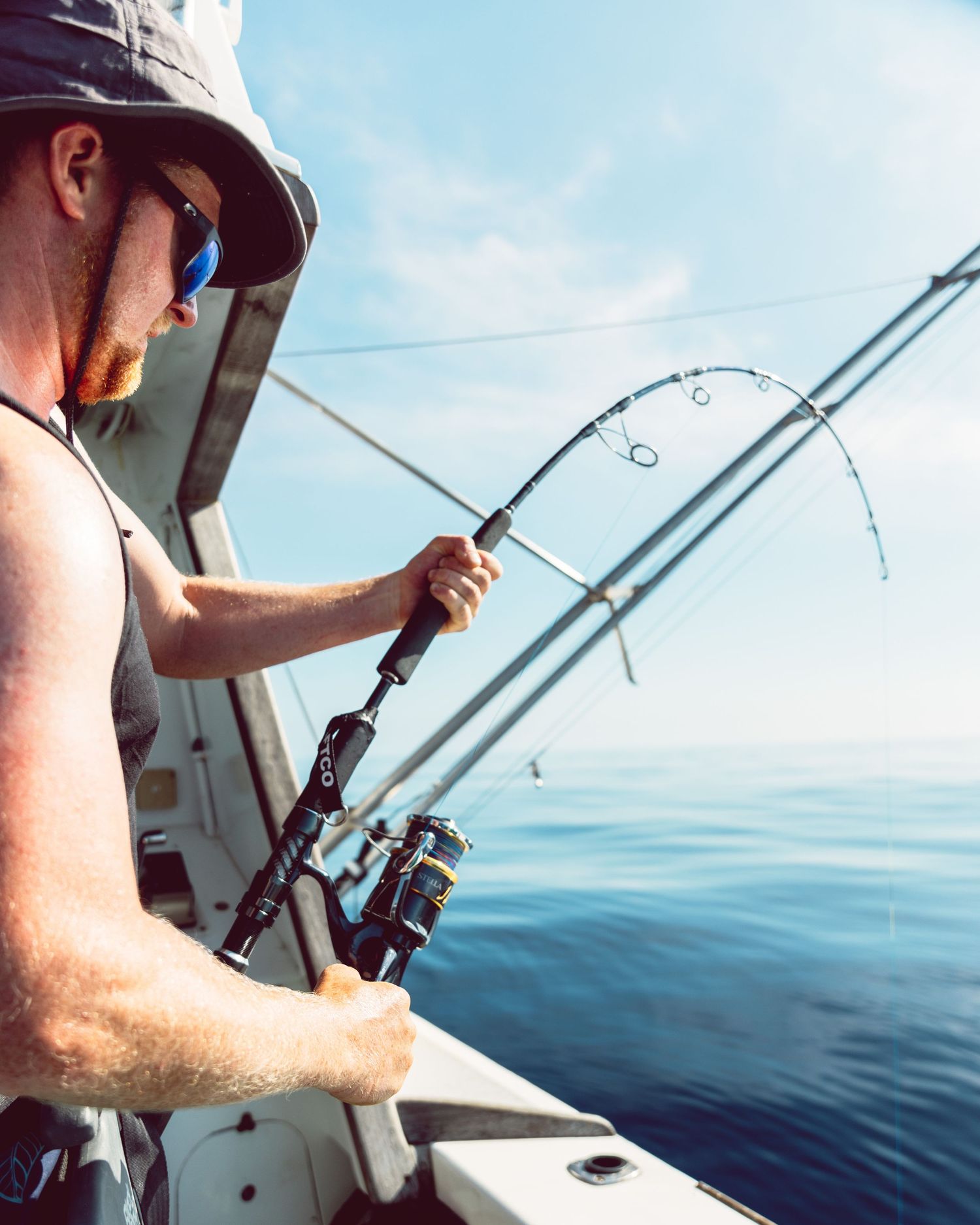 Man in bucket hat and sunglasses fishing from a boat with multiple rods over calm blue ocean waters.