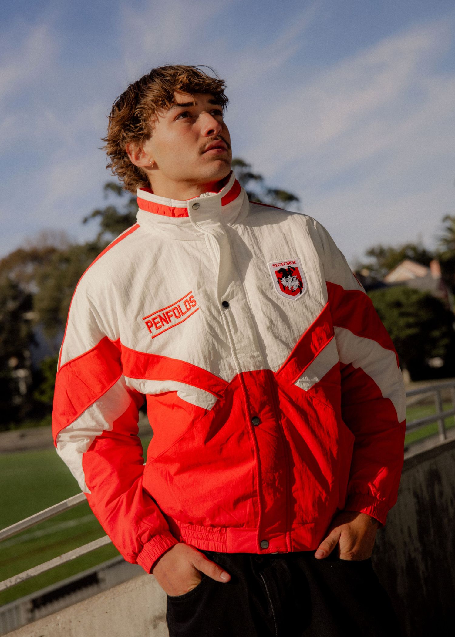 Young man wearing a red and white St George Dragons Penfolds jacket, posing outdoors near a sports field. Sutherland Shire photographer