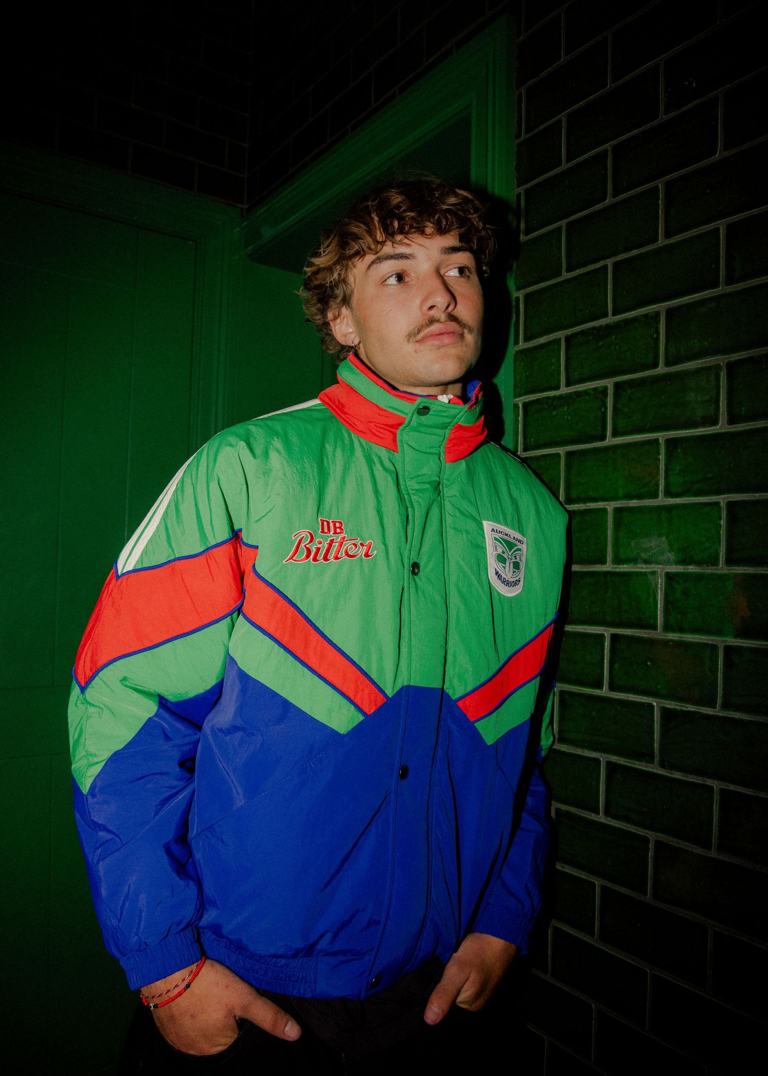 Sutherland - Young man with curly hair and mustache wearing a green, blue, and red retro sports jacket leaning against a green brick wall.