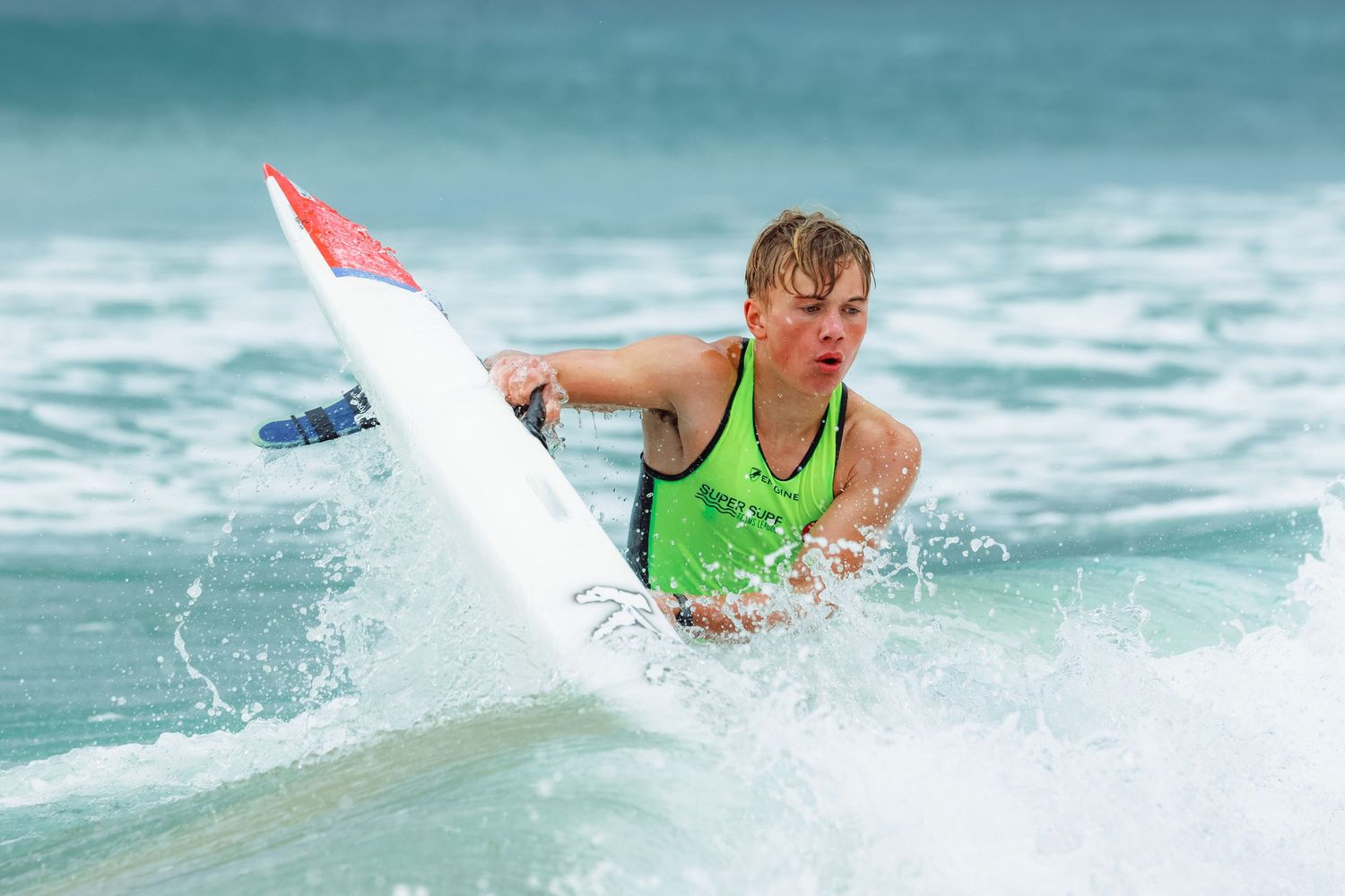 Young surfer in green vest riding a wave on a white surfboard in choppy ocean waters.