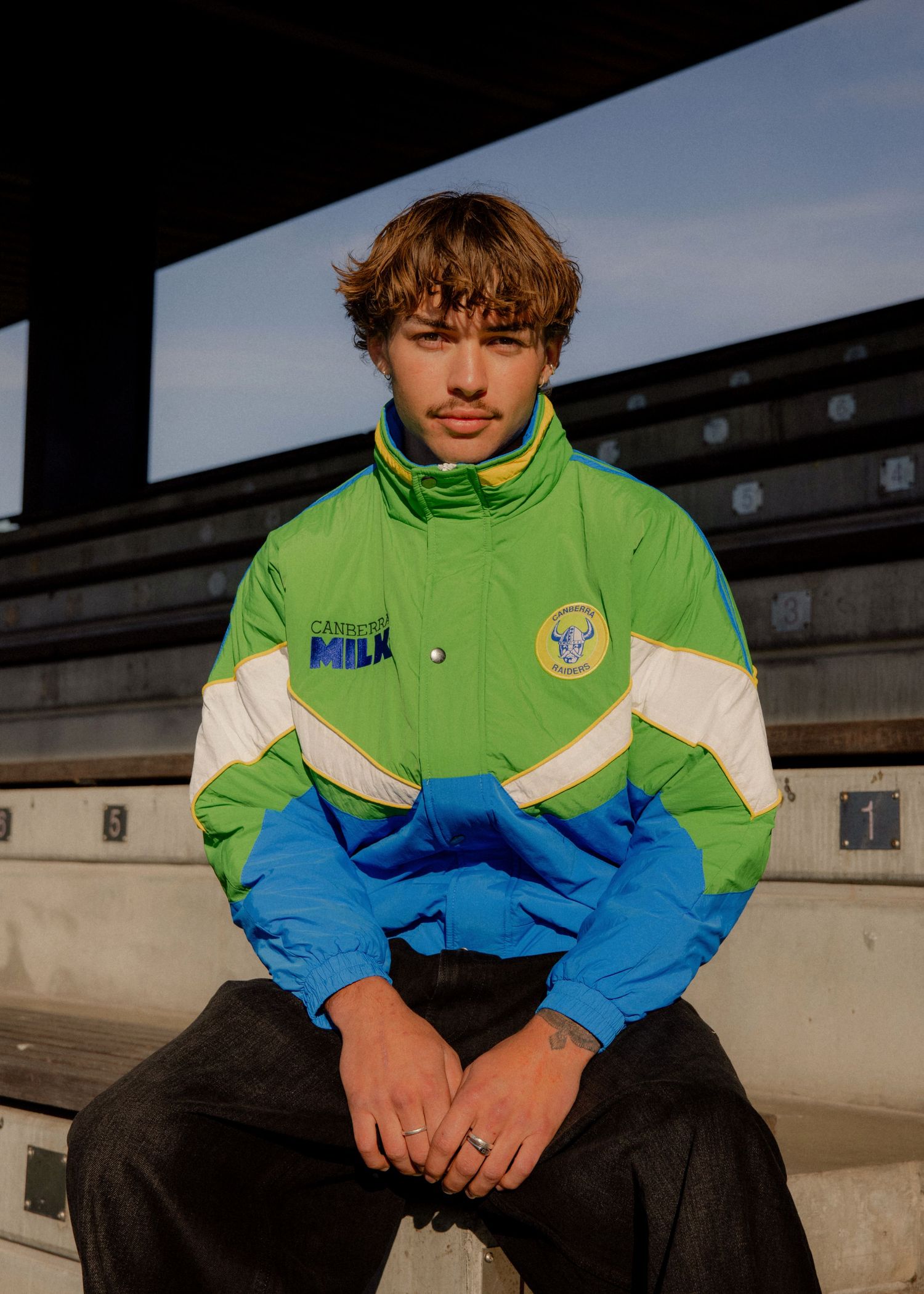 Young man sitting on bleachers wearing a retro green, white, and blue sports jacket with Milk branding. Sutherland Shire photographer