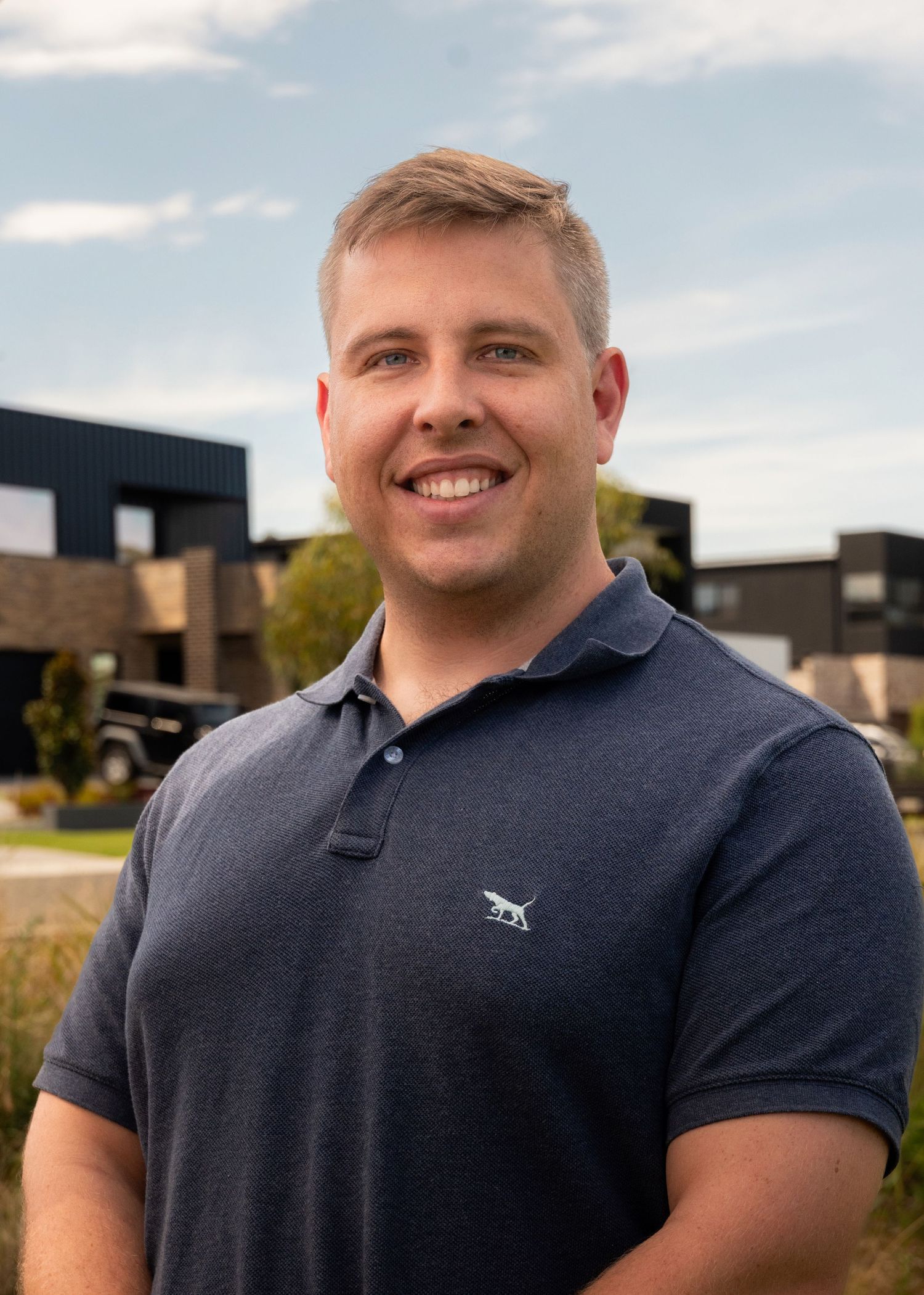 Smiling man in navy polo shirt standing outdoors near modern dark buildings on a sunny day.