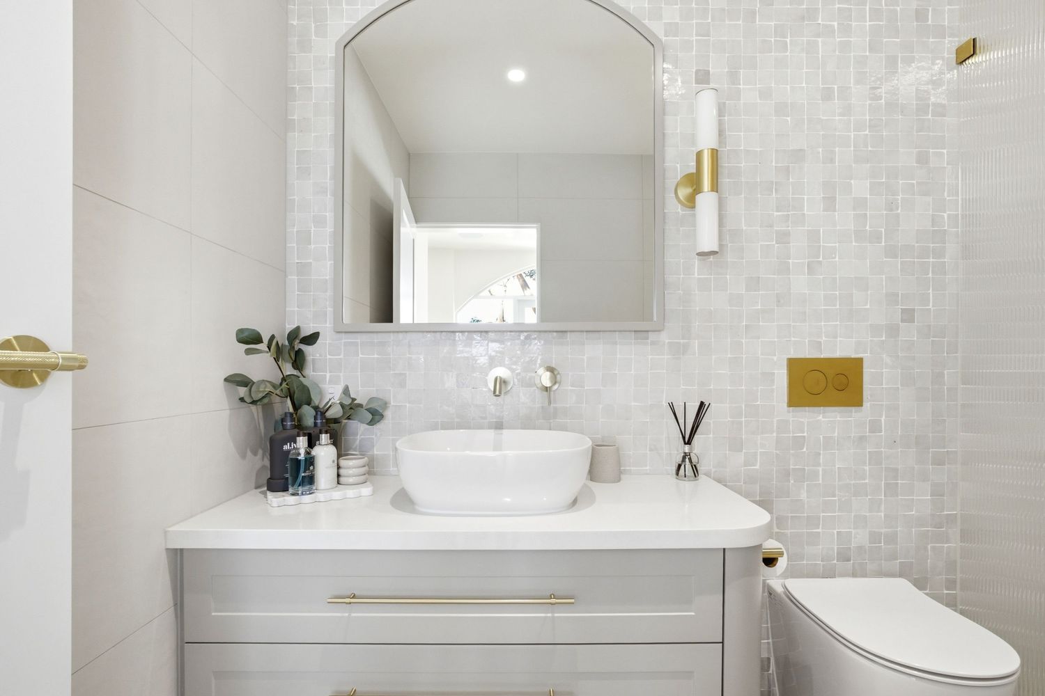 Modern white bathroom vanity with vessel sink, gold fixtures, arched mirror, and mosaic tile wall.
