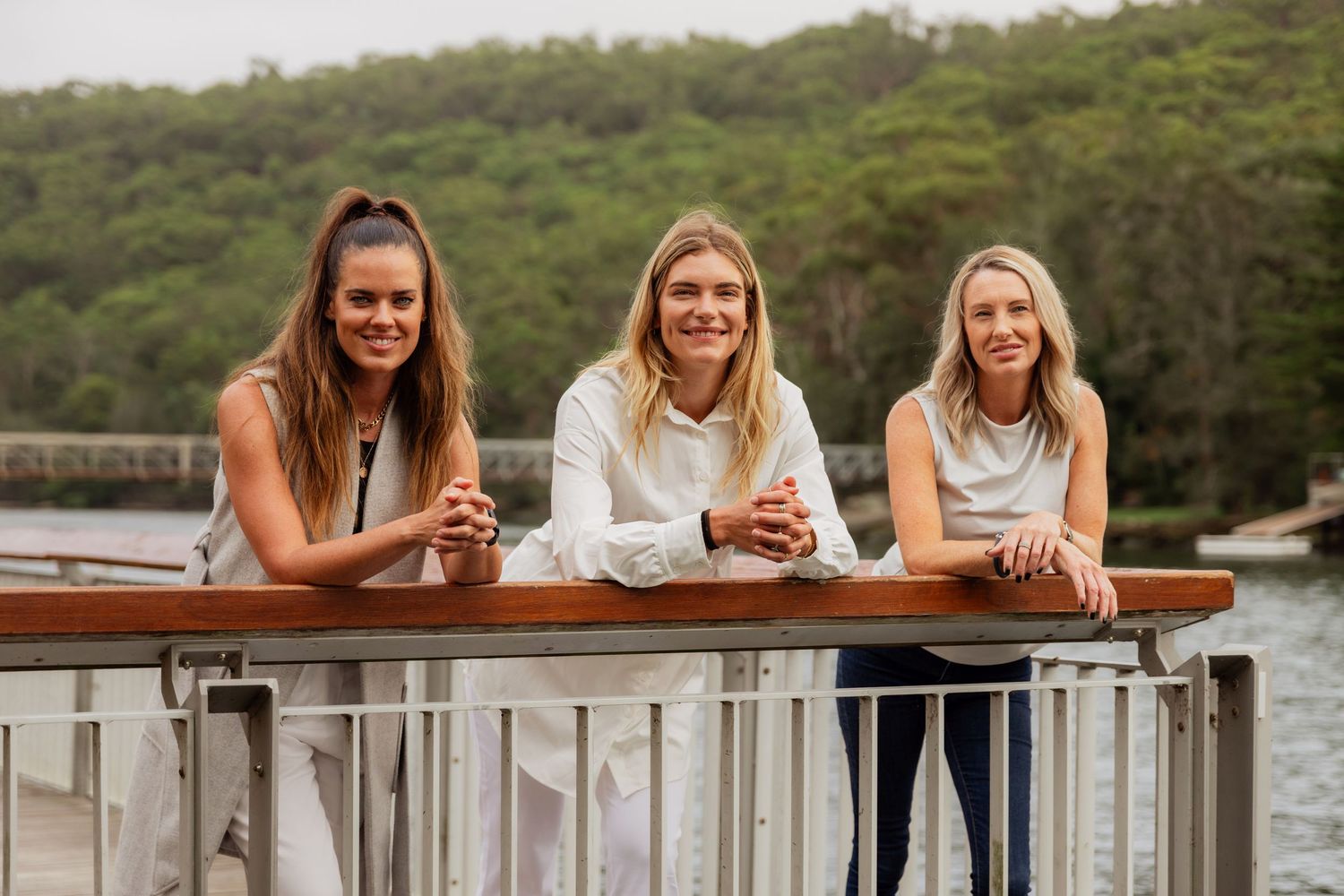 Three smiling women lean on a railing on a waterfront dock surrounded by lush green hills.