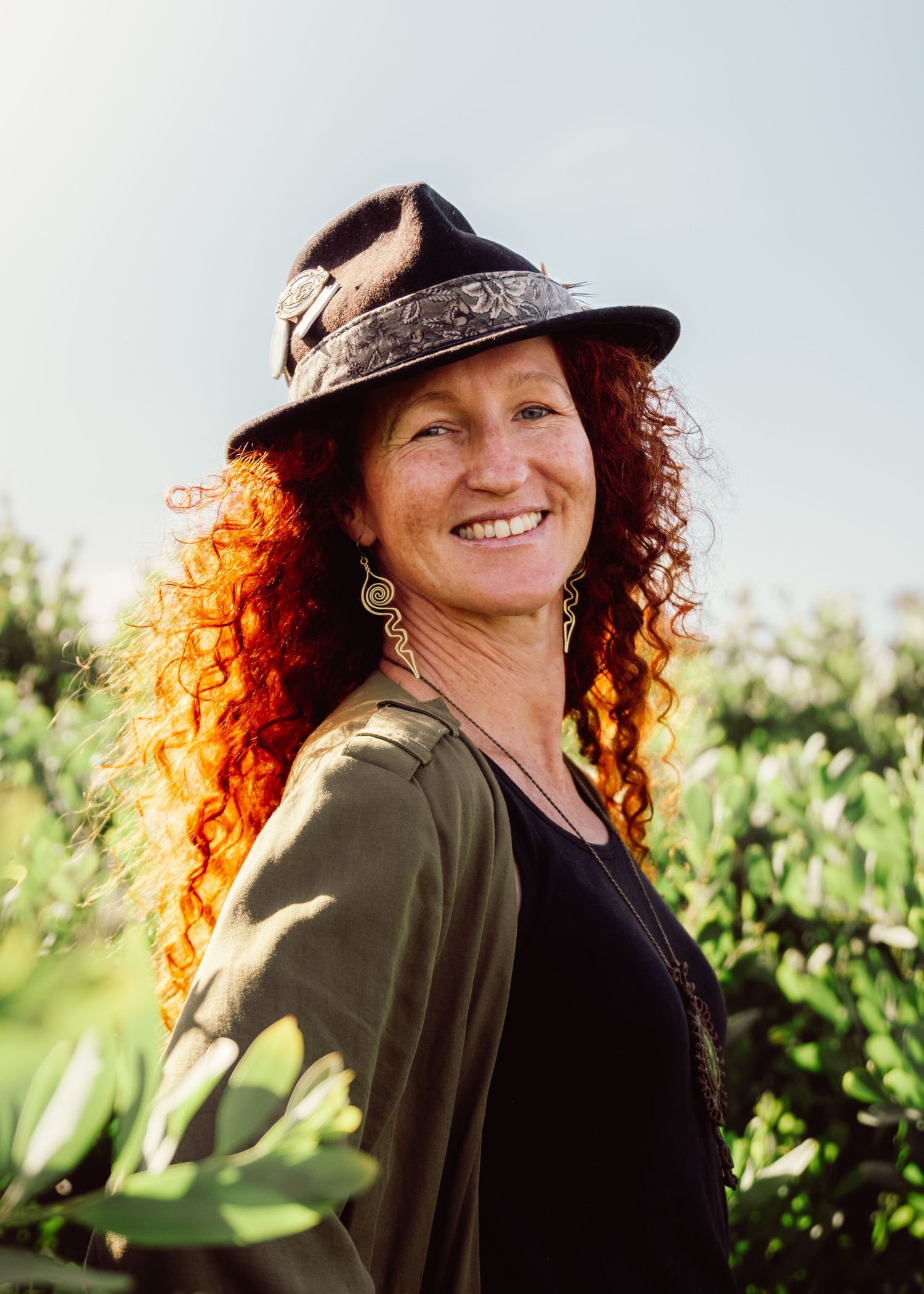 Smiling woman with curly red hair wearing a black fedora hat and green cardigan outdoors.