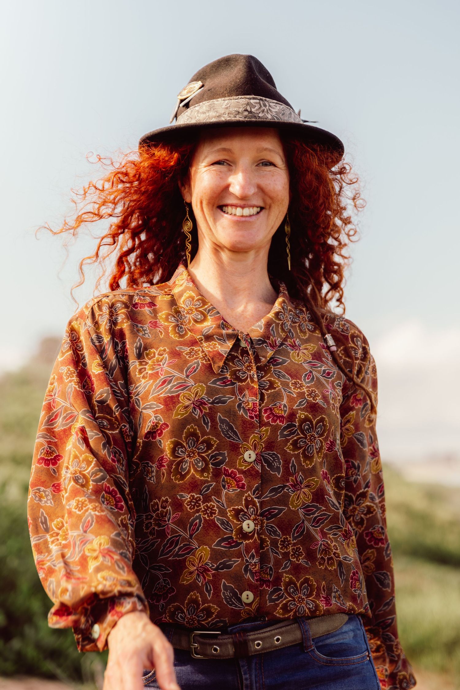 Smiling woman with red curly hair wearing a floral brown blouse and black hat outdoors.