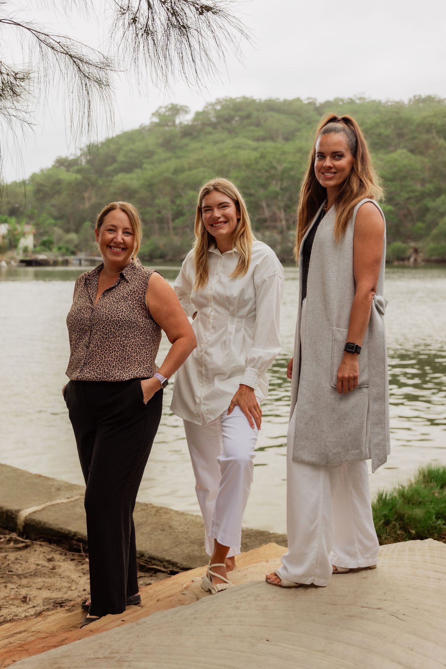 Three women smiling and posing together on a waterfront path surrounded by lush greenery.
