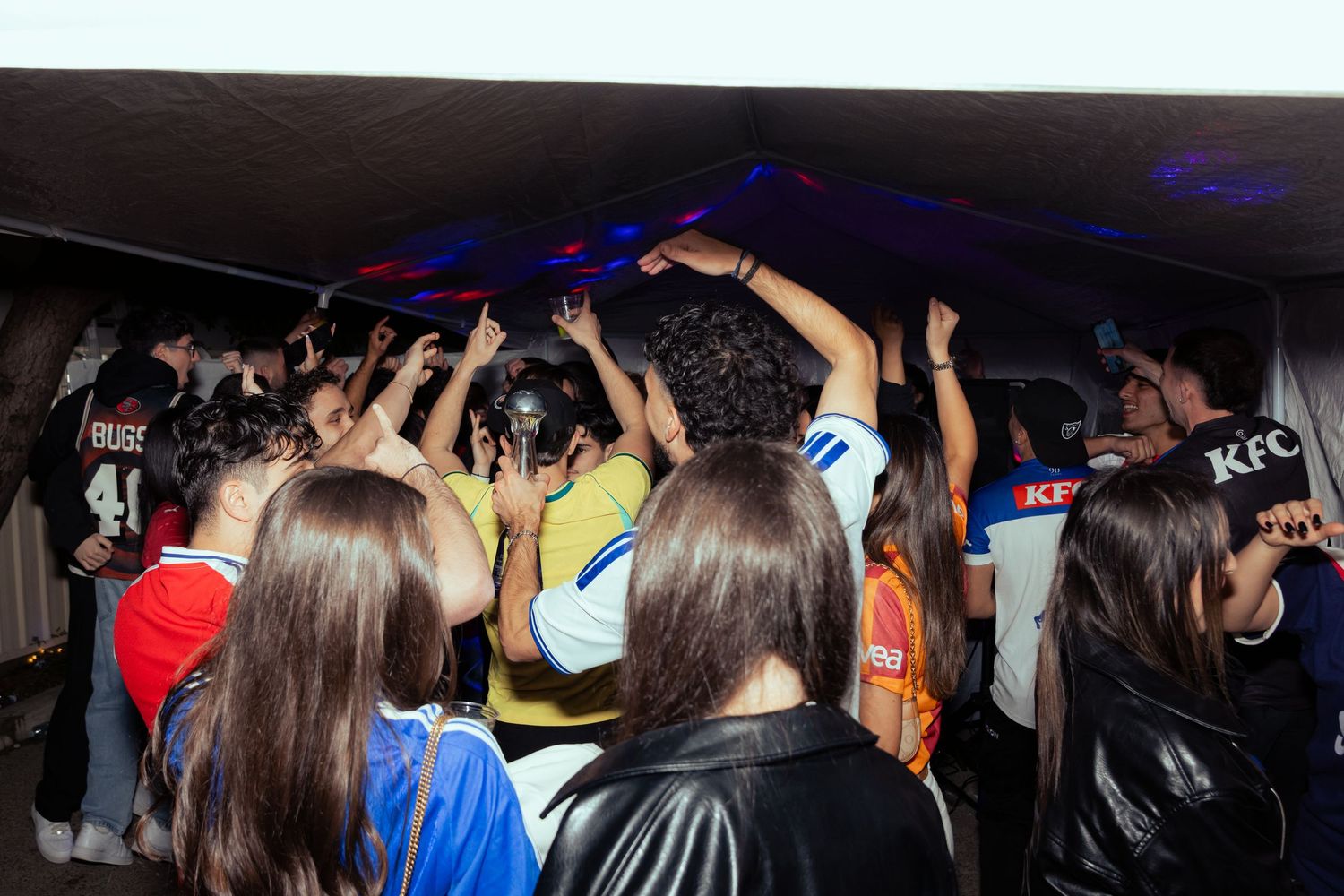 Crowd of sports fans dancing and celebrating under a tent with blue lights at a lively fan event.