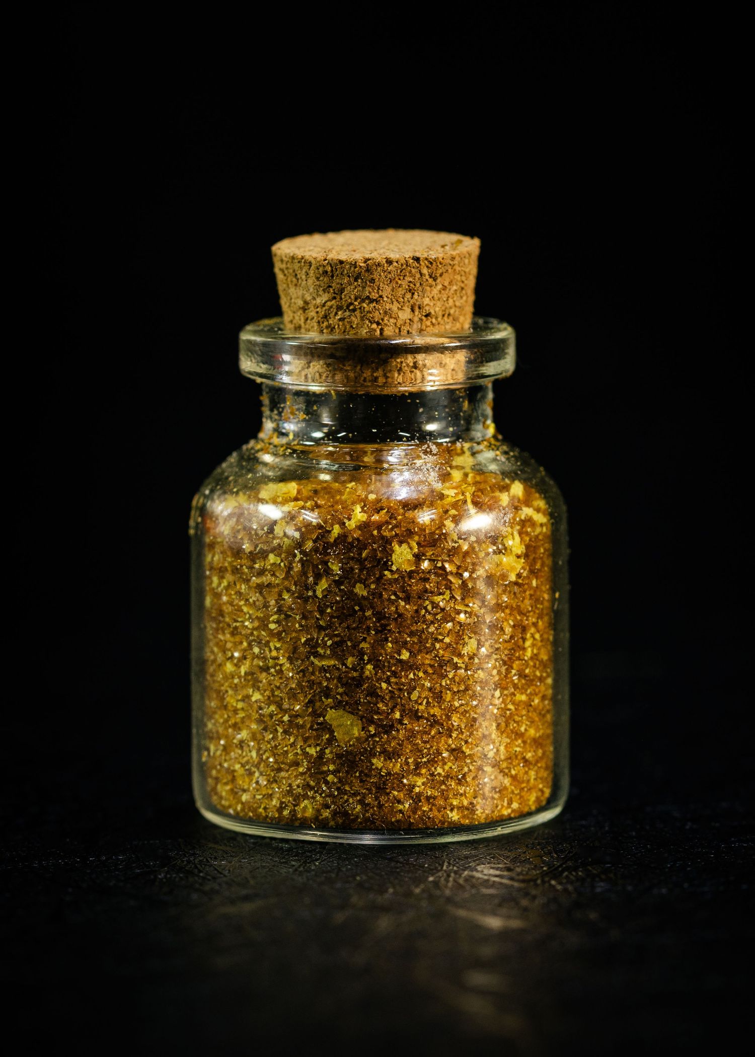 Small glass bottle with cork stopper filled with golden glitter and gold flakes on a dark background.