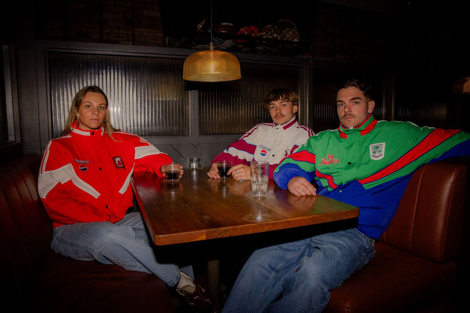 Three people in colorful retro sportswear sit together at a wooden table in a dimly lit bar. Business and brand photographer