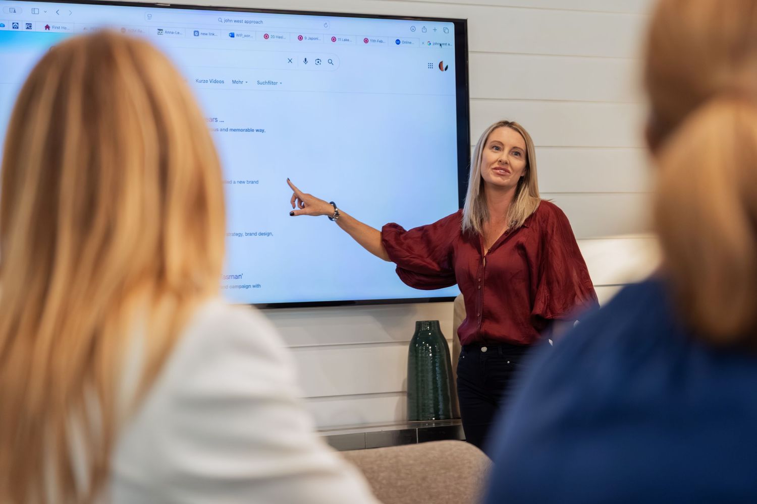 A woman in a red blouse presents at a digital whiteboard during a business meeting with colleagues.