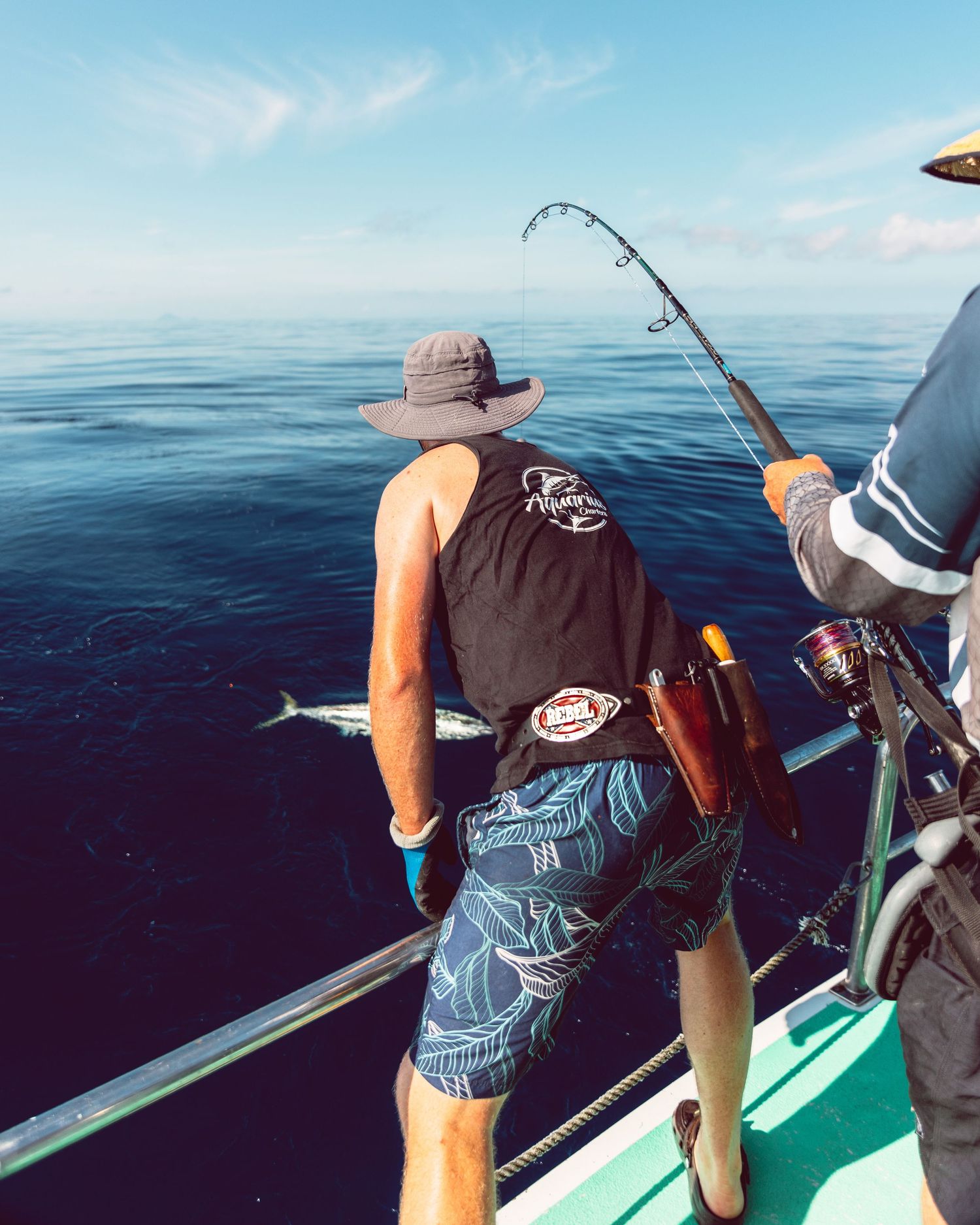 Two anglers fishing from a boat on calm blue ocean waters, one holding a bent rod with a fish on the line.