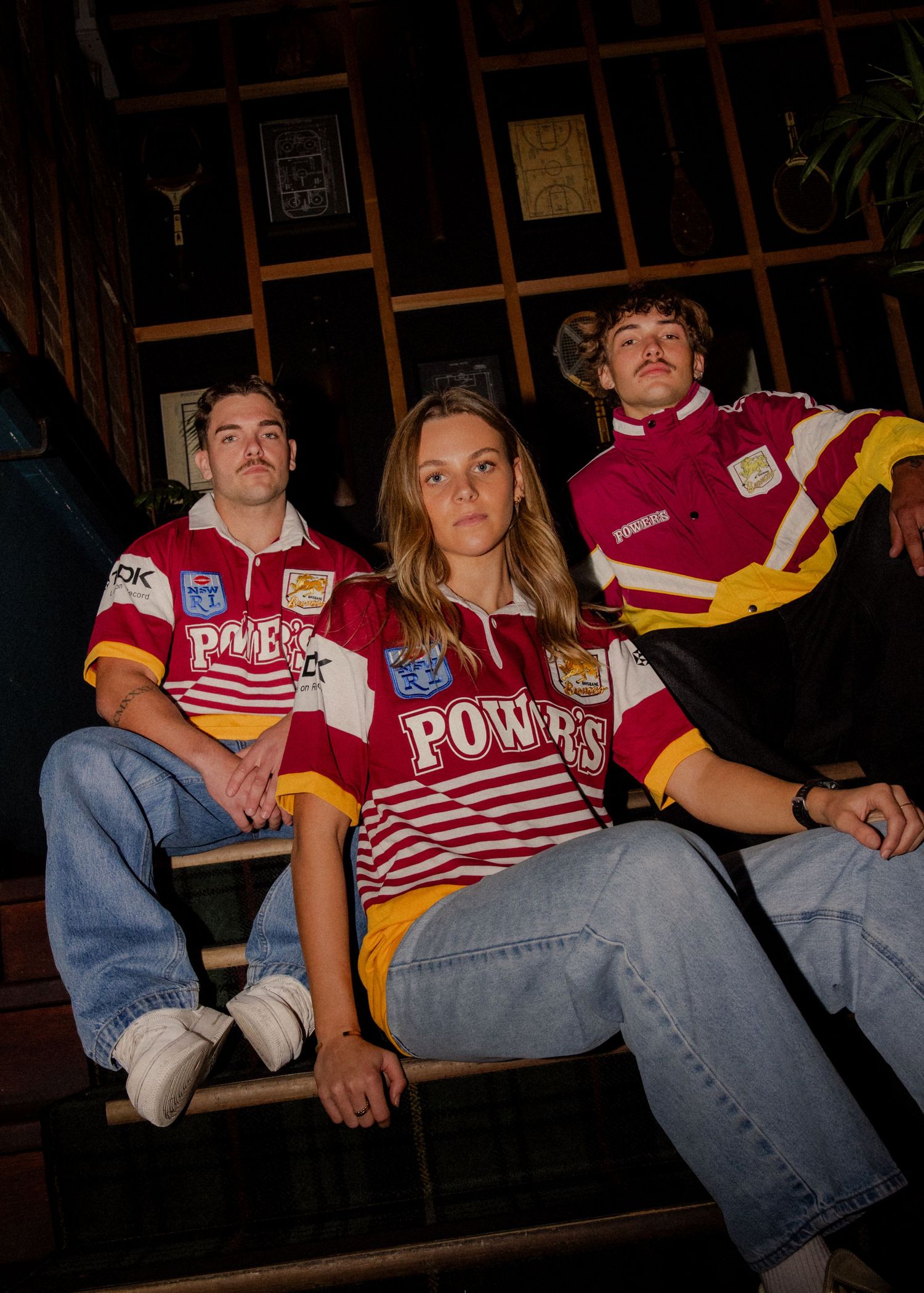 Three young adults wearing vintage red and gold striped Powers rugby jerseys, seated on wooden stairs indoors. Sutherland Shire photographer