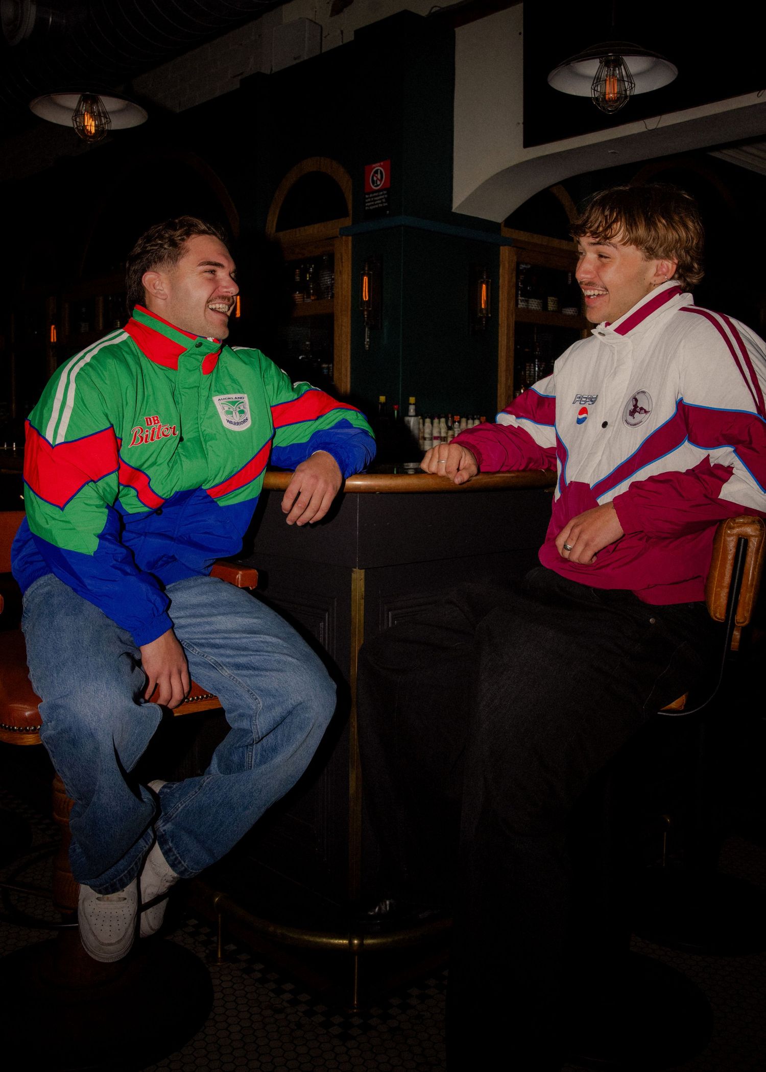 Two men in retro NRL rugby league jackets laugh together on bar stools in a dimly lit pub. Business and brand photographer sydney
