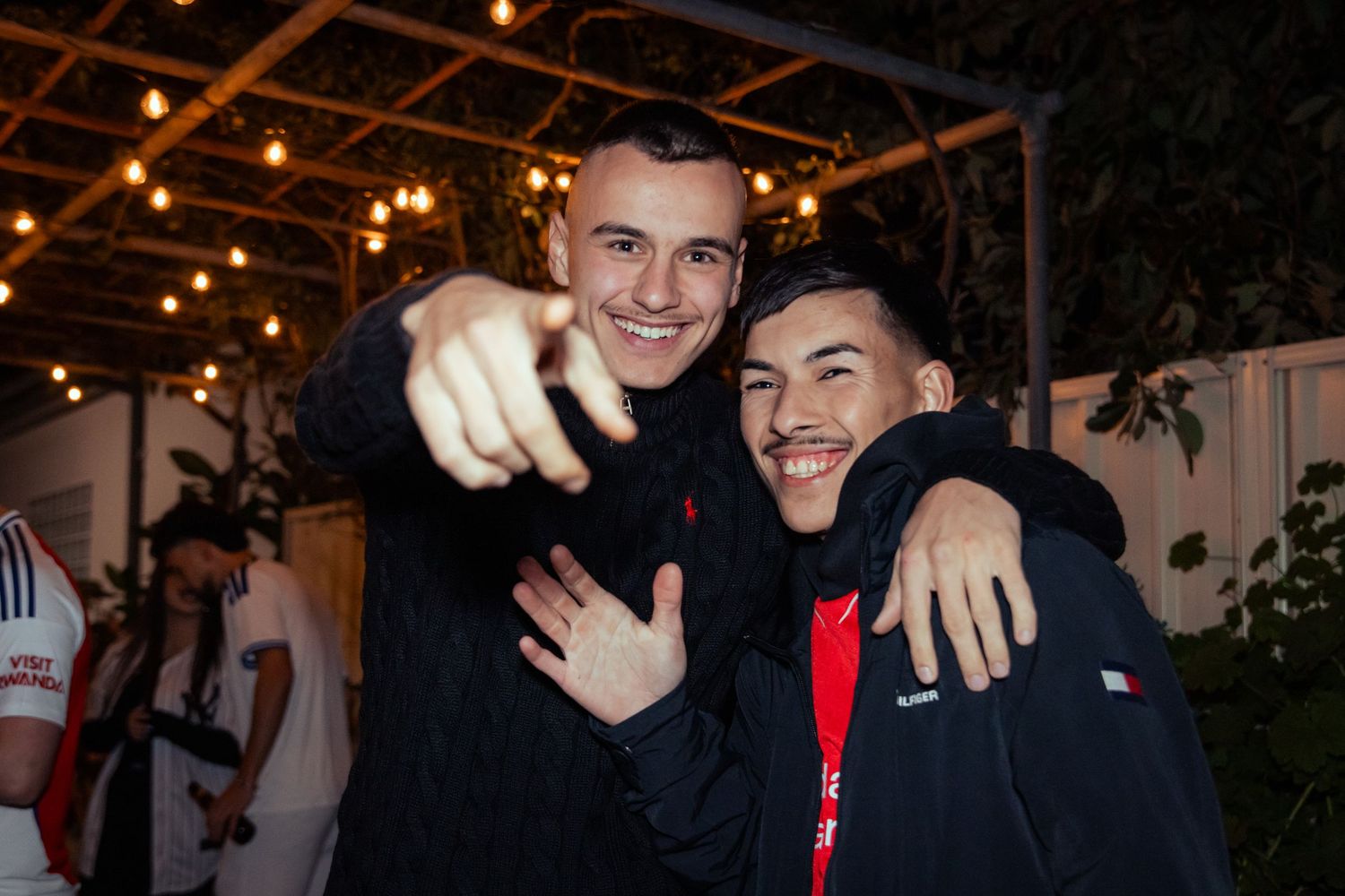 Two young men in black jackets smiling and pointing at camera at an outdoor evening event.