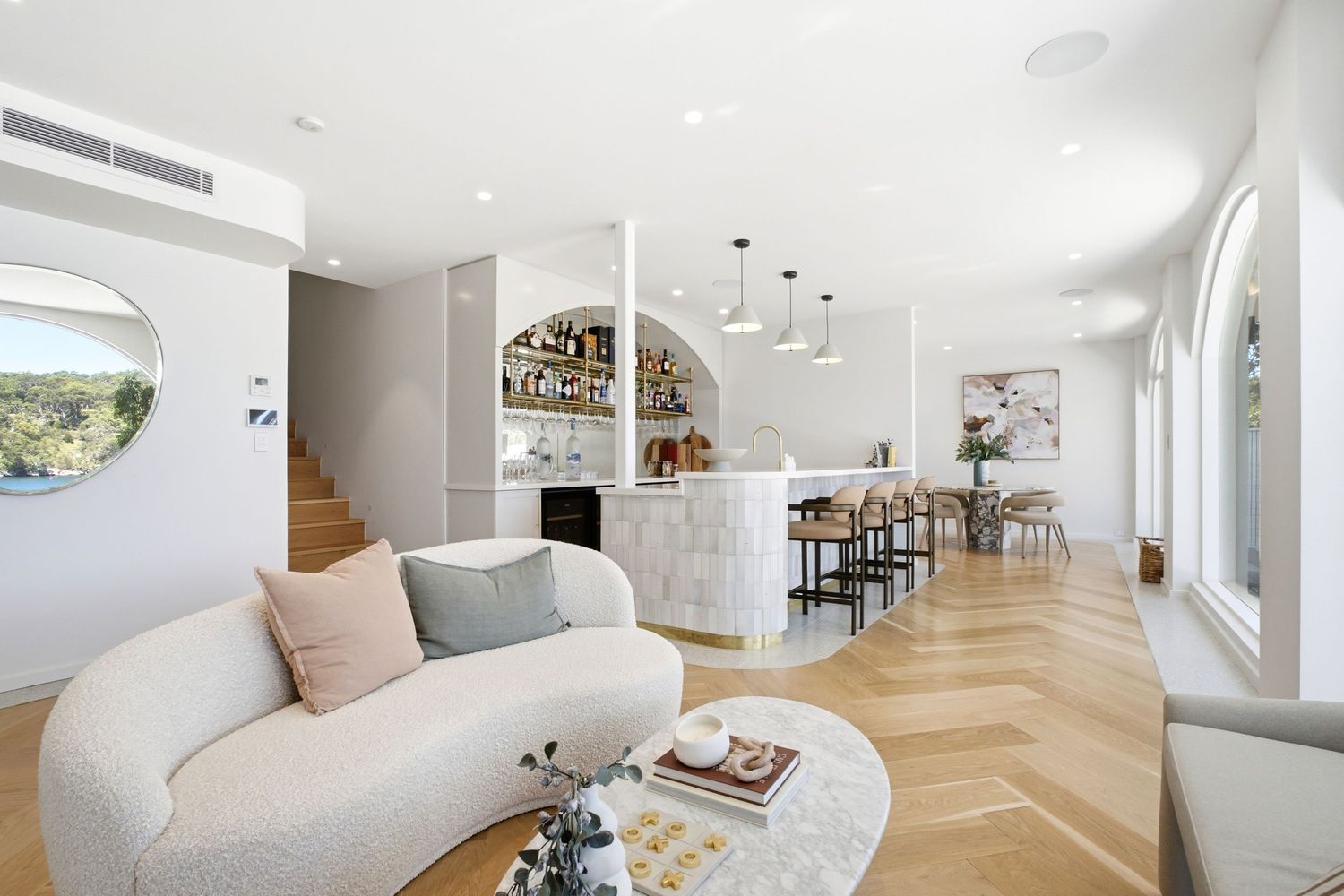 Elegant open-plan living area with curved sofa, marble bar, pendant lights, herringbone timber floors, and arched windows.