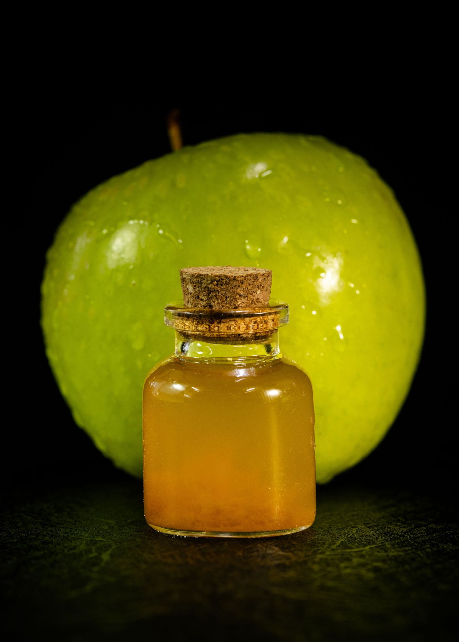 Small glass bottle with cork filled with apple cider vinegar beside a fresh green apple on dark background.