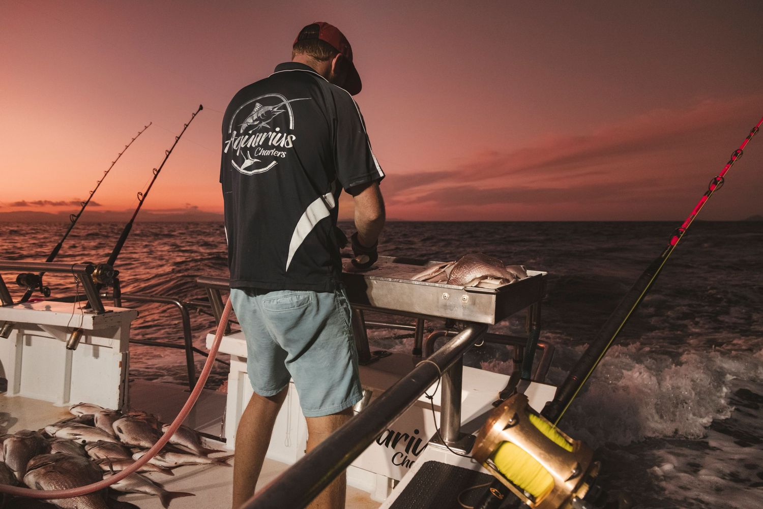 Fisherman on a boat at sunset with fishing rods, wearing a black shirt, ocean waves in the background.
