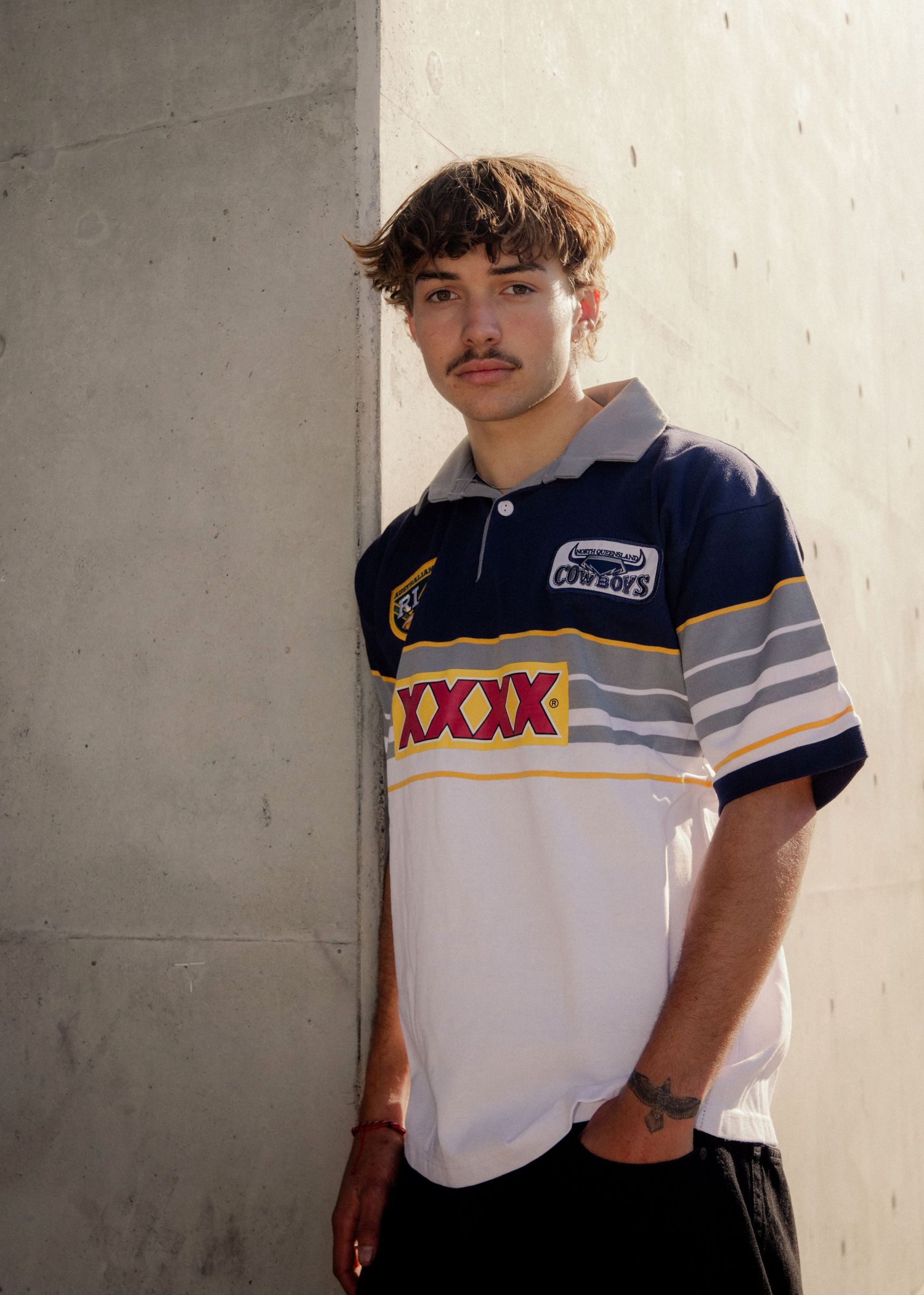 Young man with mustache wearing a XXXX Cowboys rugby league jersey leaning against a concrete wall.