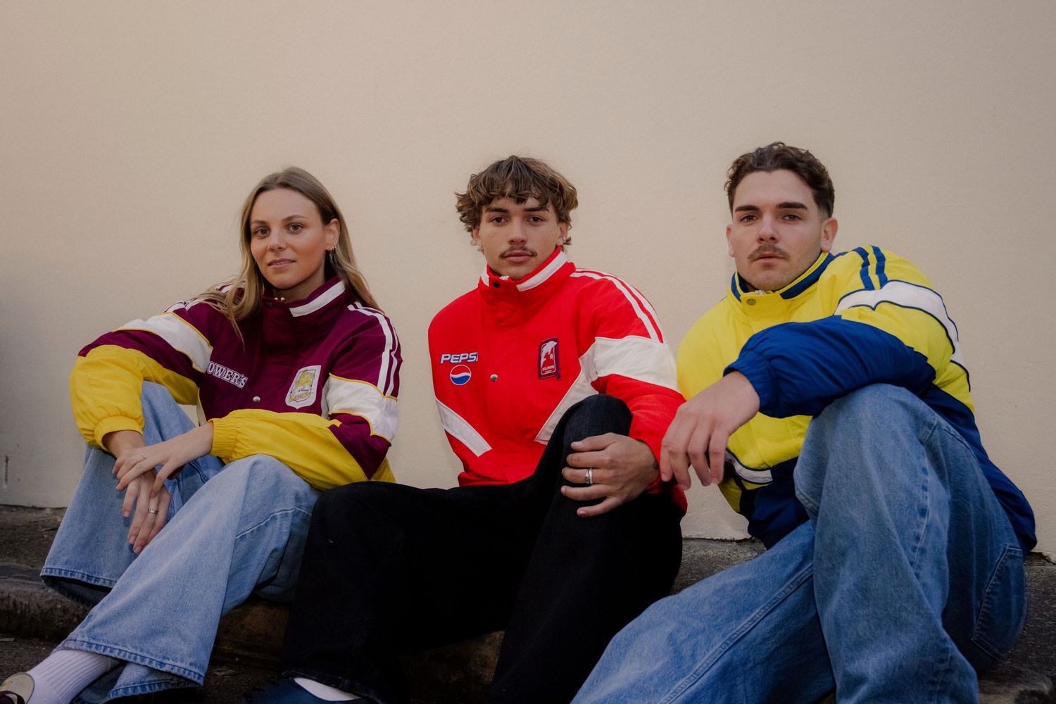 Three models sitting outdoors wearing retro NRL rugby league tracksuit jackets in maroon, red and yellow.