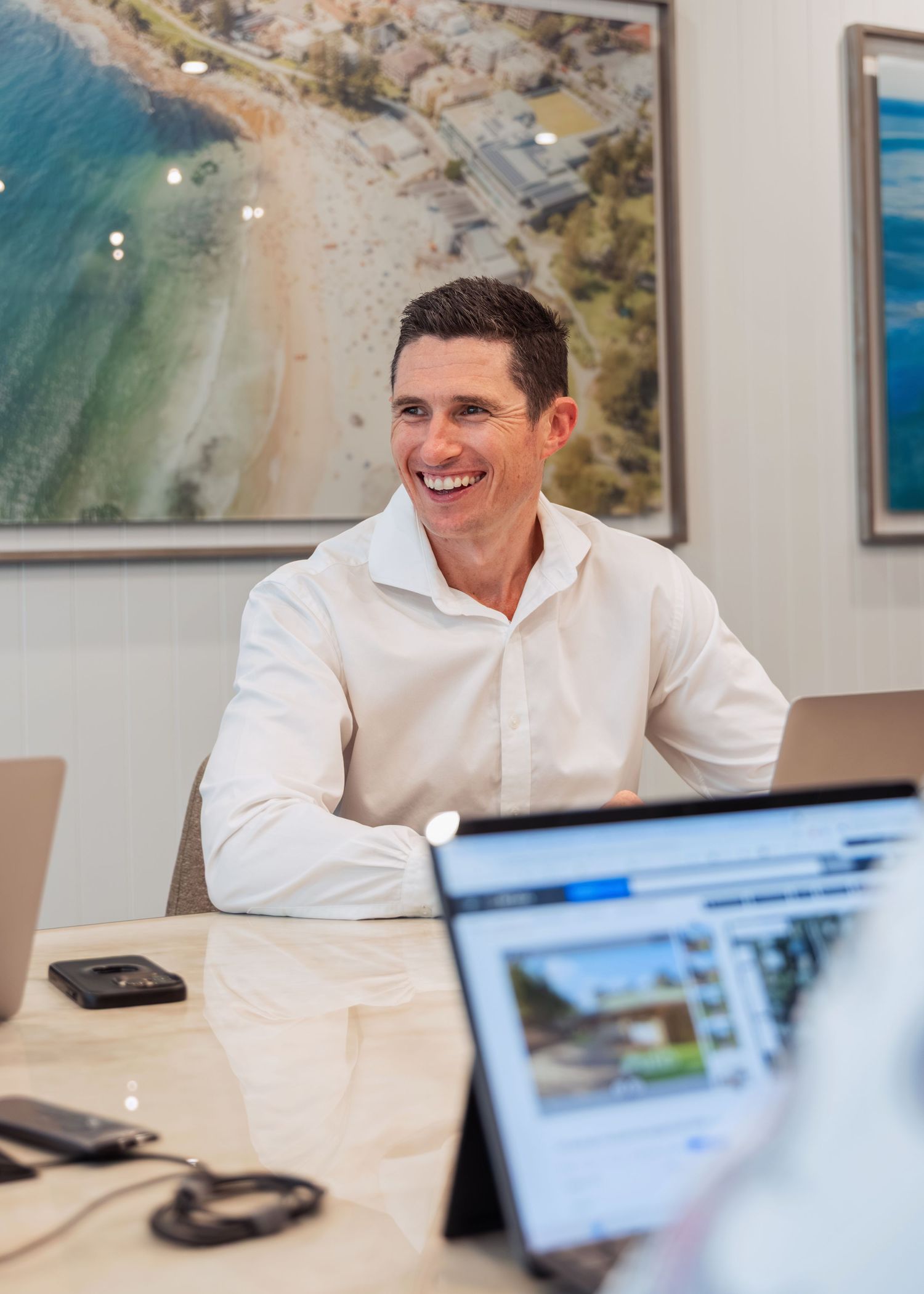 Smiling man in white shirt sitting at conference table with aerial beach photo on wall behind him.