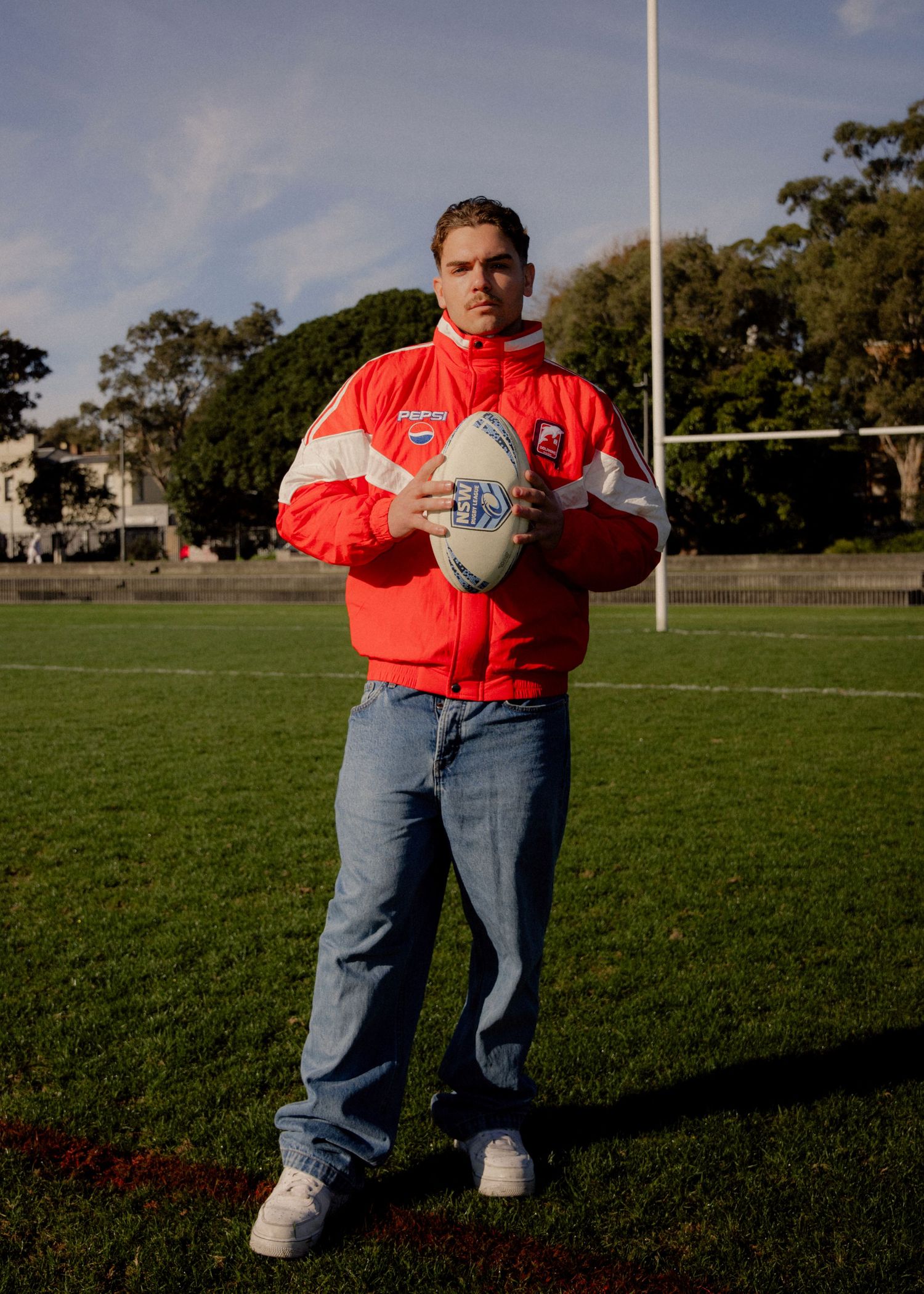 Man in red sports jacket holding rugby league ball on green field near goal posts. Business and brand photographer sydney
