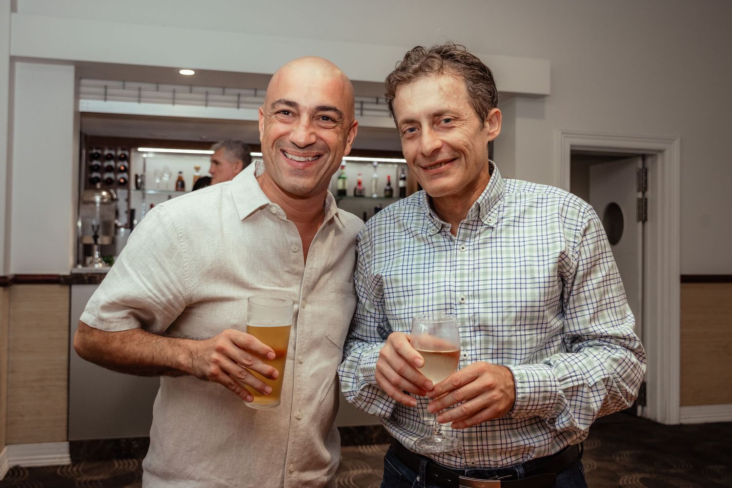 Two smiling men holding drinks at a casual indoor social gathering or bar event.