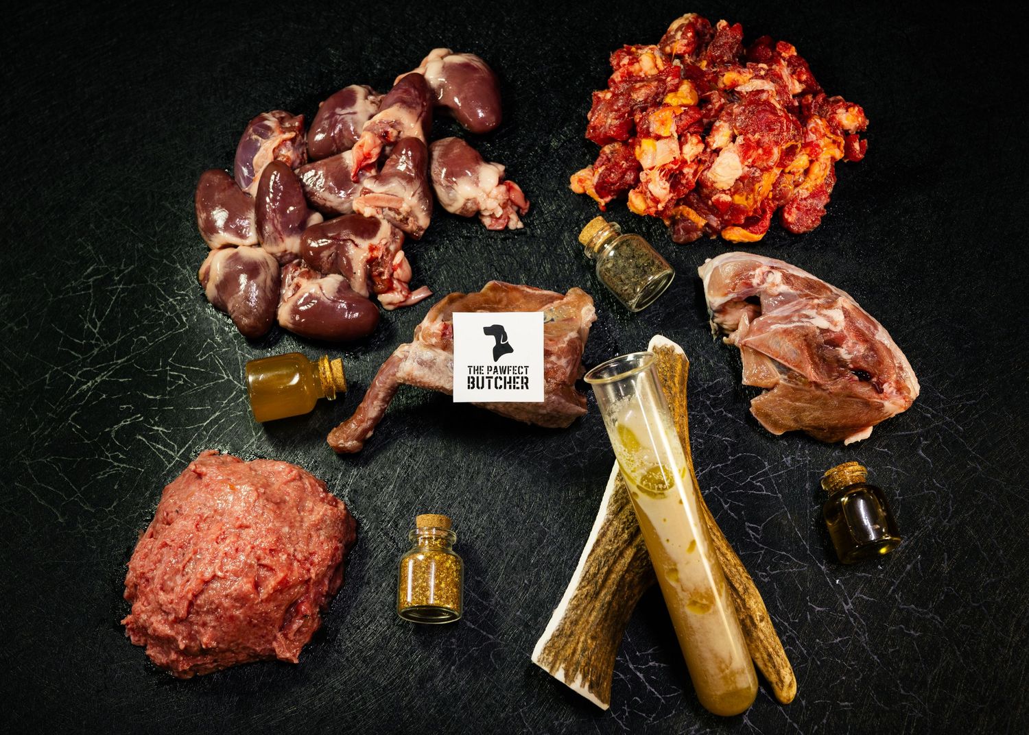 Assorted raw dog food ingredients including organ meats, ground beef, bones, and supplements on a dark background.