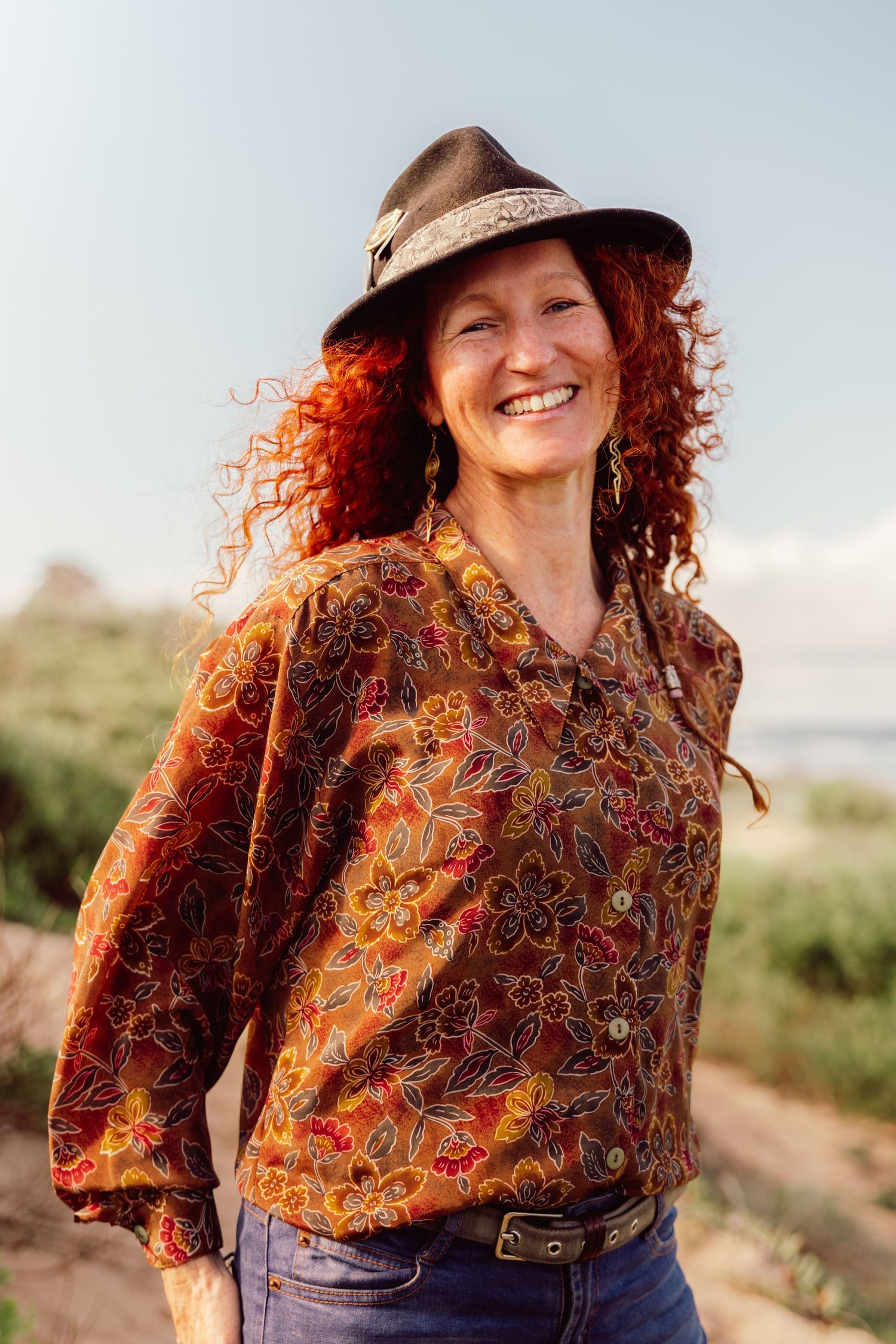 Smiling woman with curly red hair wearing a floral blouse, fedora hat, and jeans outdoors.