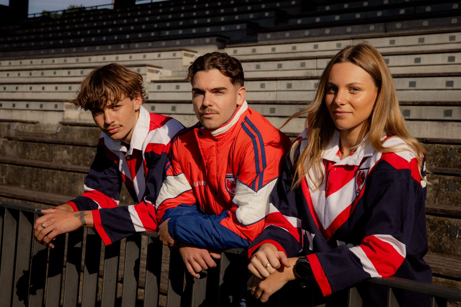 Three young people wearing matching red, white, and blue retro track jackets sitting on stadium bleachers outdoors. Business and brand photo