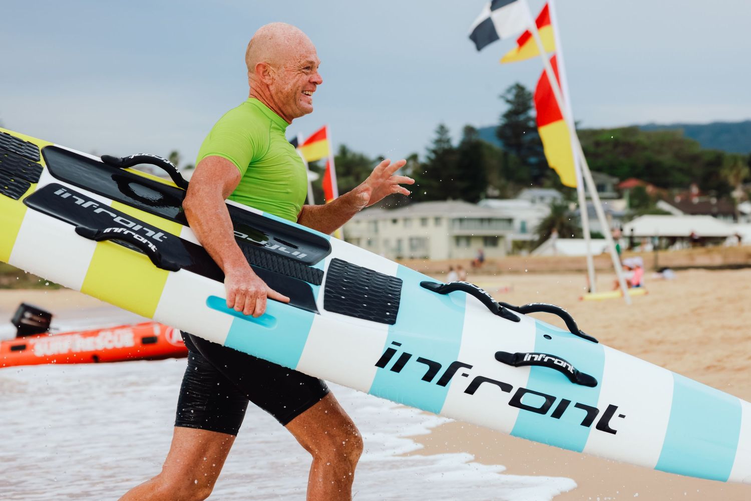 Man in green shirt carrying an Infront paddleboard on a sunny beach with colorful flags in the background.