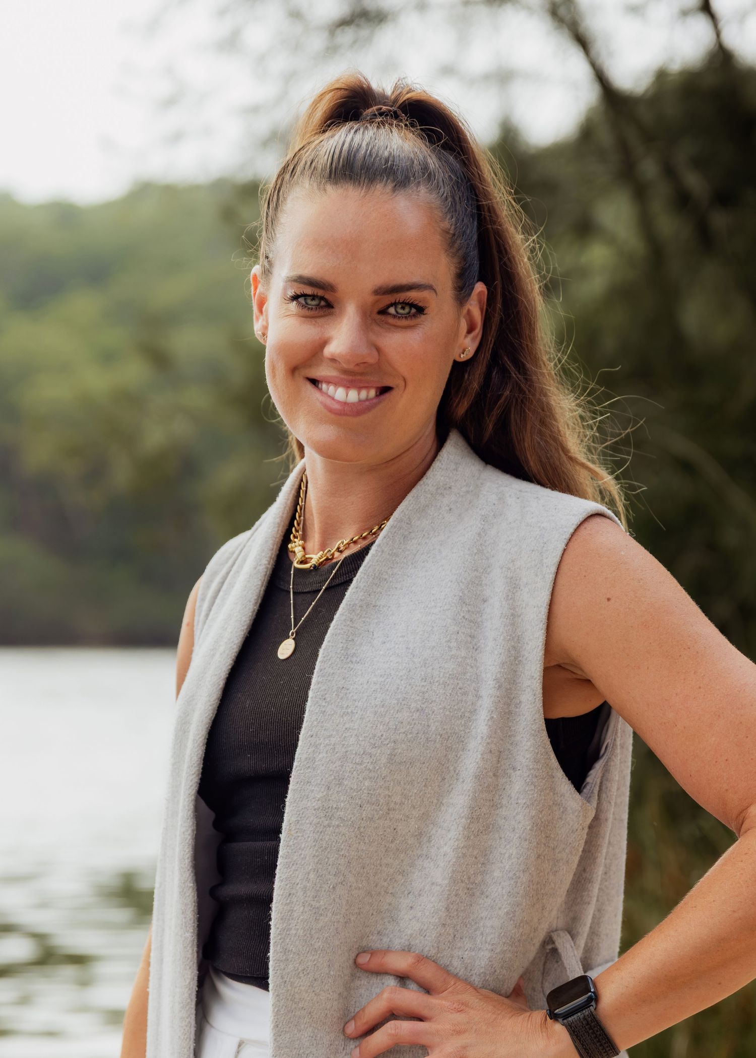 Smiling woman with ponytail wearing gray vest and gold necklaces poses outdoors near a lake with trees.