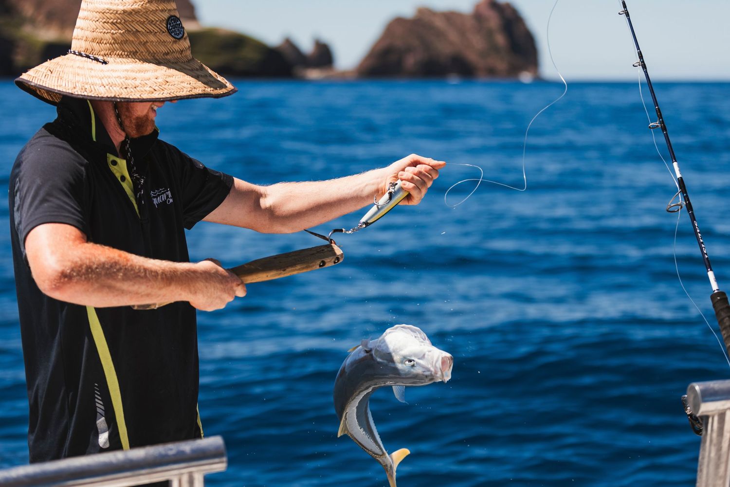 Man in straw hat fishing on a boat, holding a caught fish over the ocean railing with a fishing rod nearby.