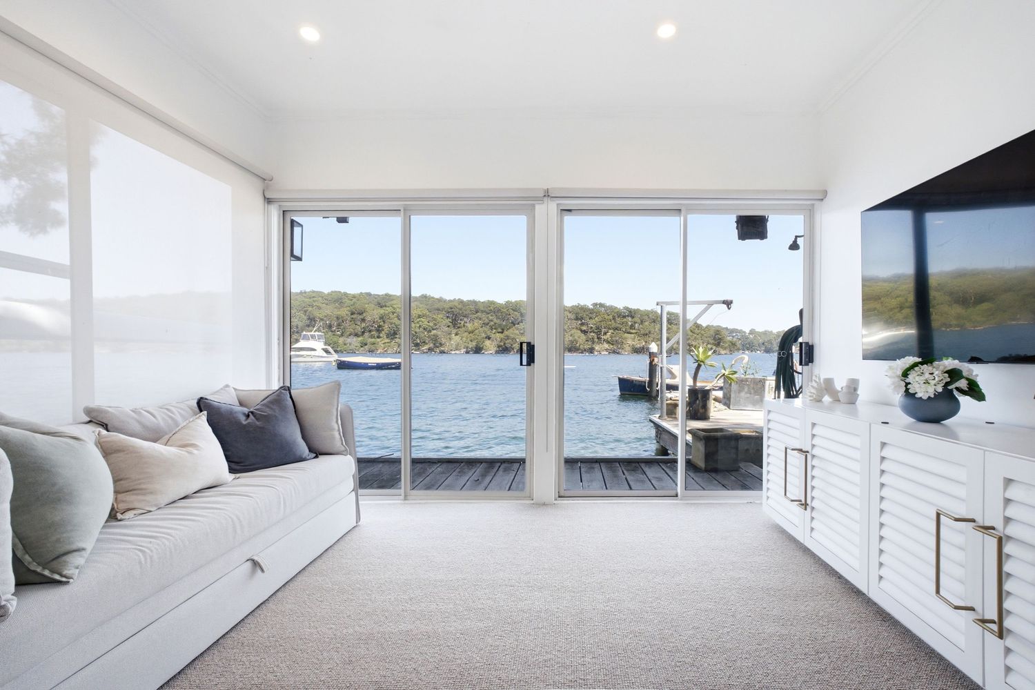 Bright living room with floor-to-ceiling sliding doors overlooking a serene waterfront deck and lush tree views.