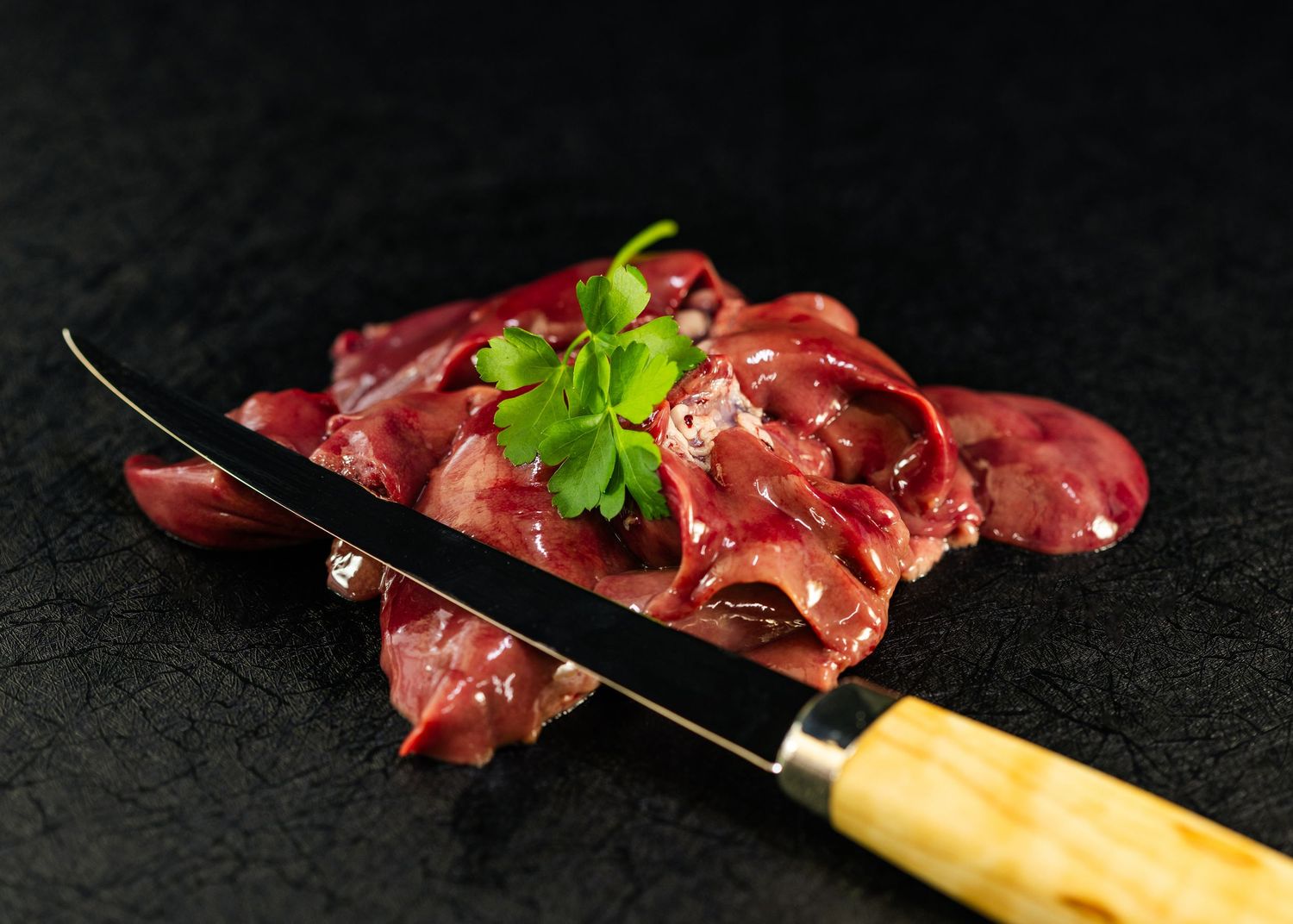 Raw chicken livers on a dark slate surface garnished with fresh parsley, with a wooden-handled knife beside them.