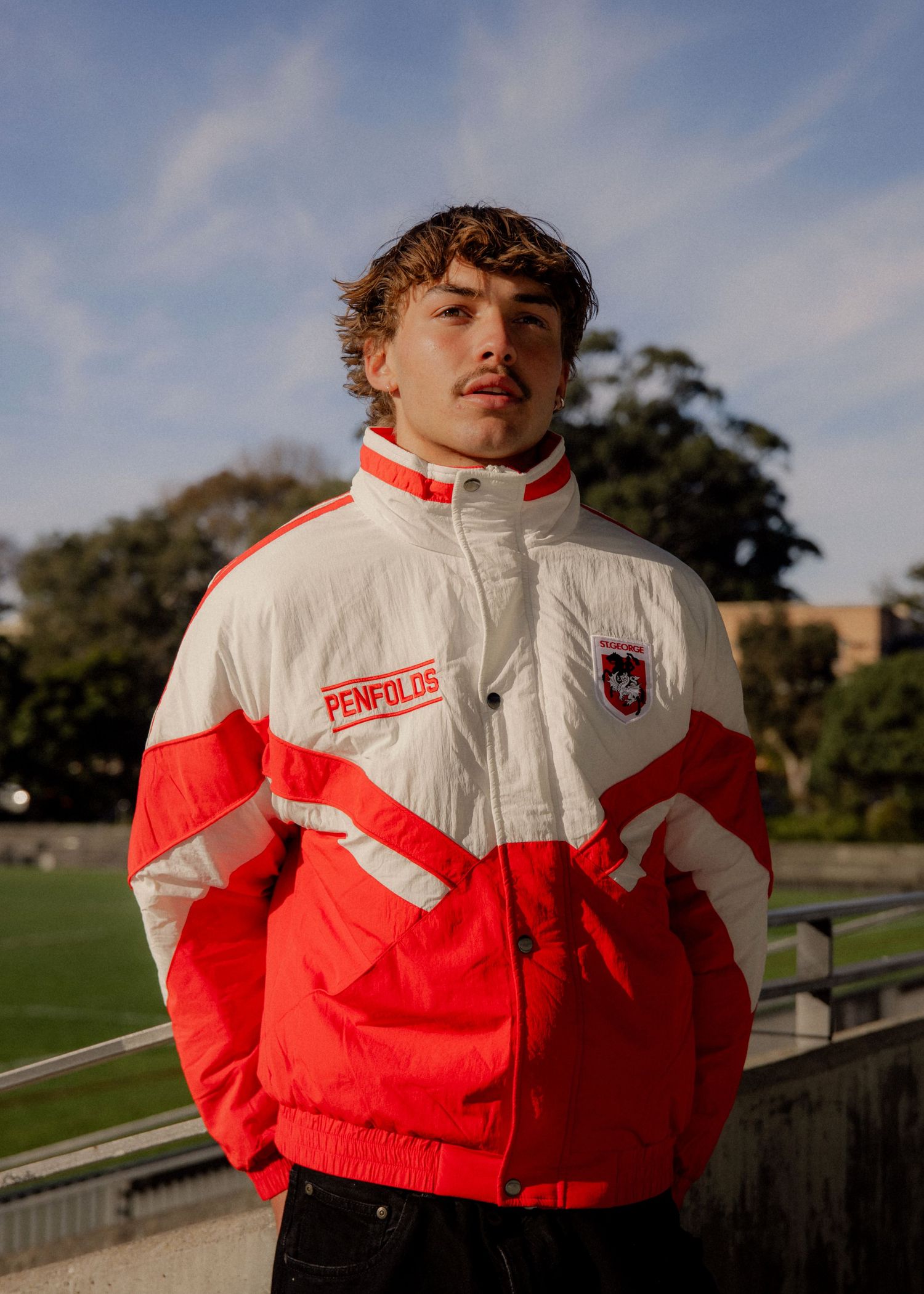 Young man wearing a red and white Penfolds St George Dragons Retro branded sports jacket standing outdoors near a sports field.