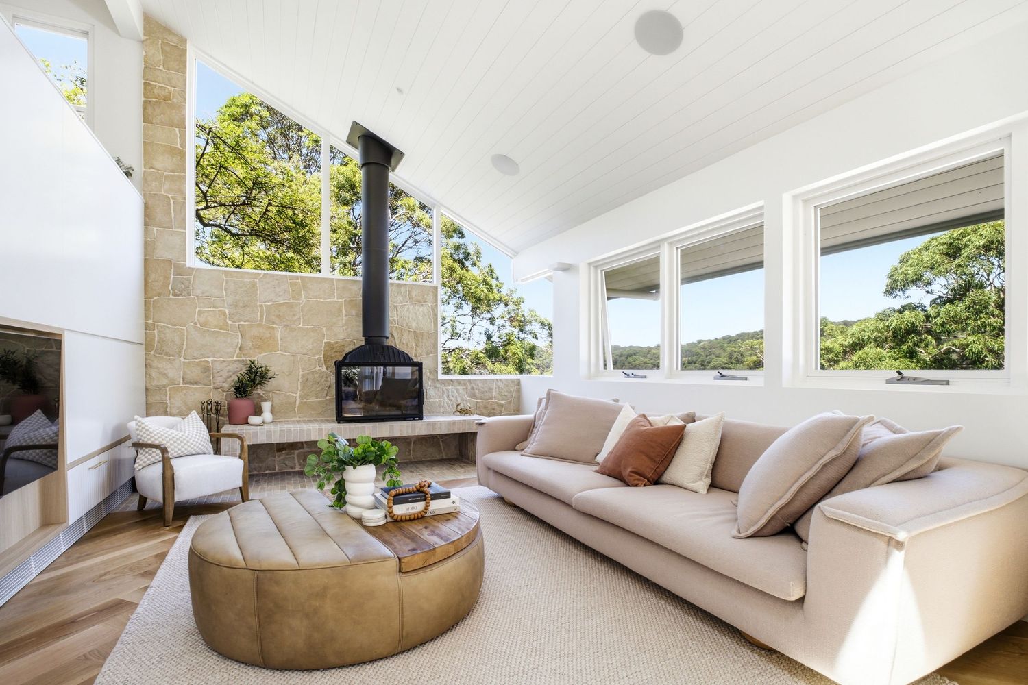 Modern living room with beige sofa, round leather coffee table, stone fireplace, and wood-burning stove with garden views.