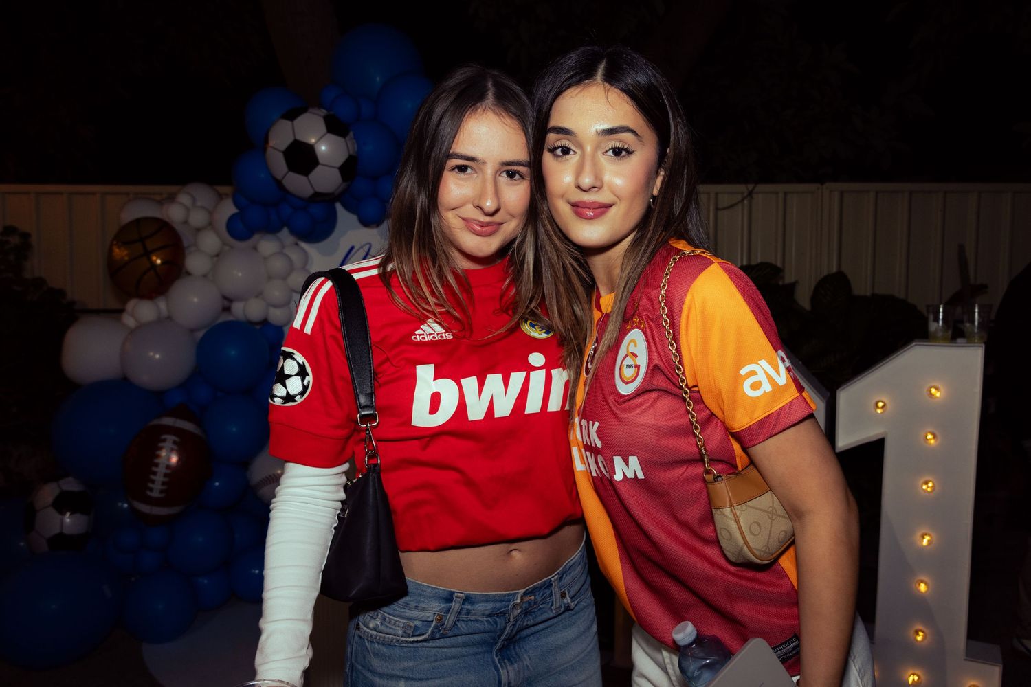 Two young women posing together wearing soccer jerseys at a sports-themed party with balloons.