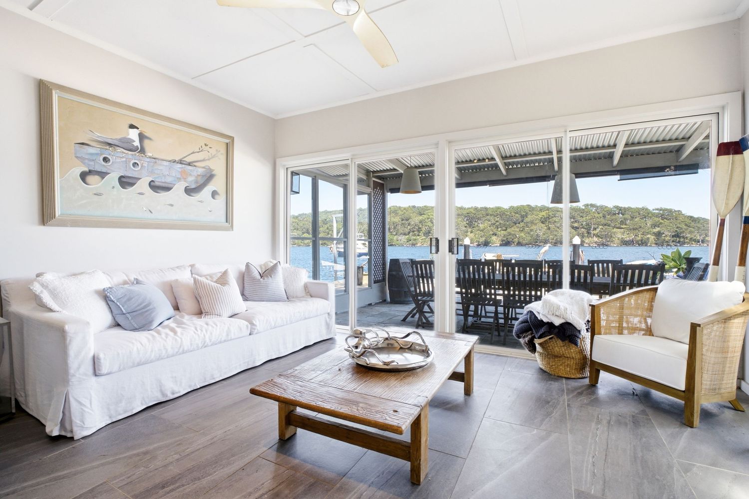 Bright coastal living room with white sofa, timber coffee table, and glass doors opening to a waterfront deck with ocean views.