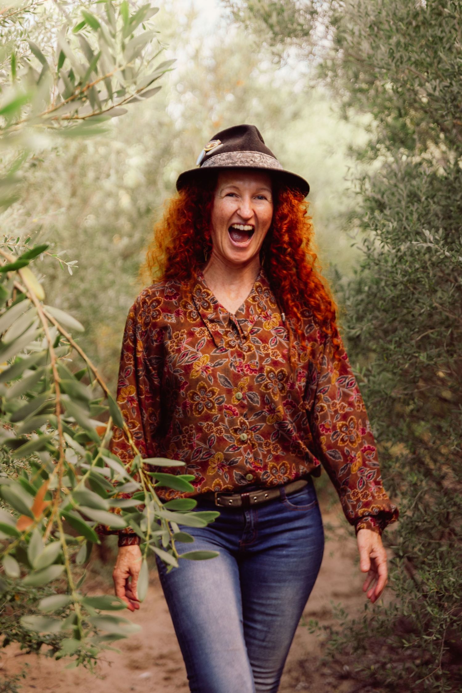 A joyful redhead woman in a floral blouse and hat laughs while walking through a lush olive grove.