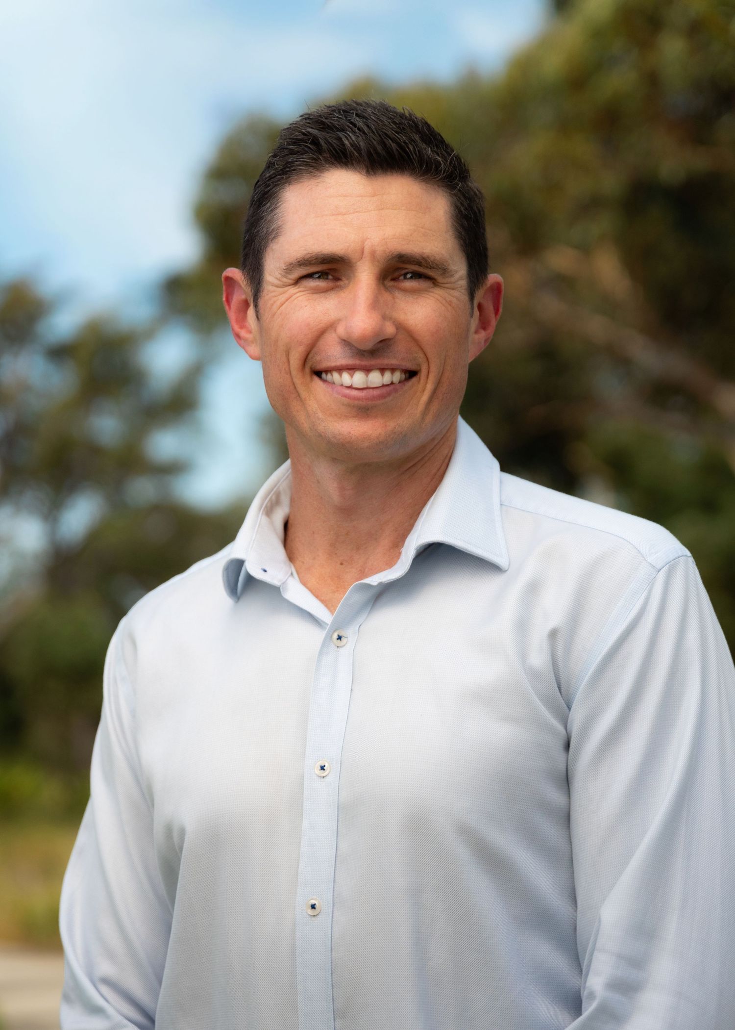 Smiling man in light blue shirt outdoors with trees in background, professional headshot photo.