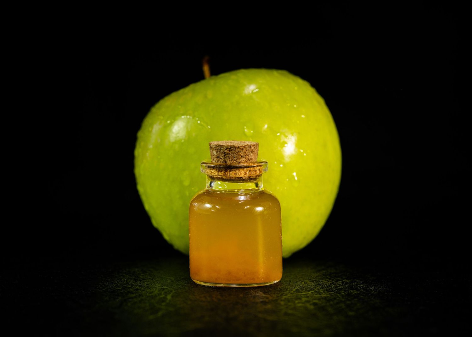 Food and product photography and videography Small glass bottle with amber liquid next to green apple on dark background.
