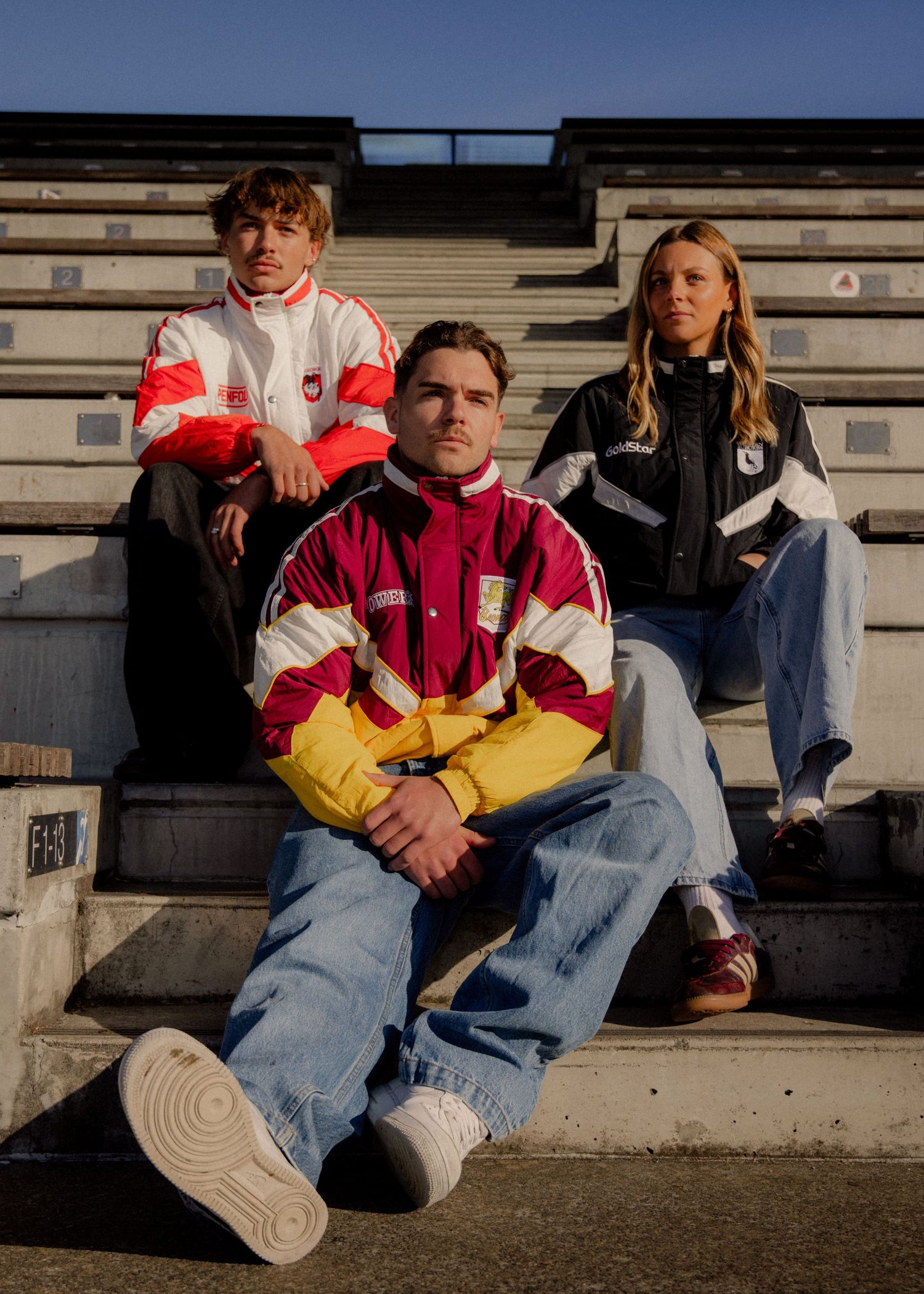 Three young people wearing retro sports jackets pose on stadium bleachers in golden hour light. Sutherland Shire videographer