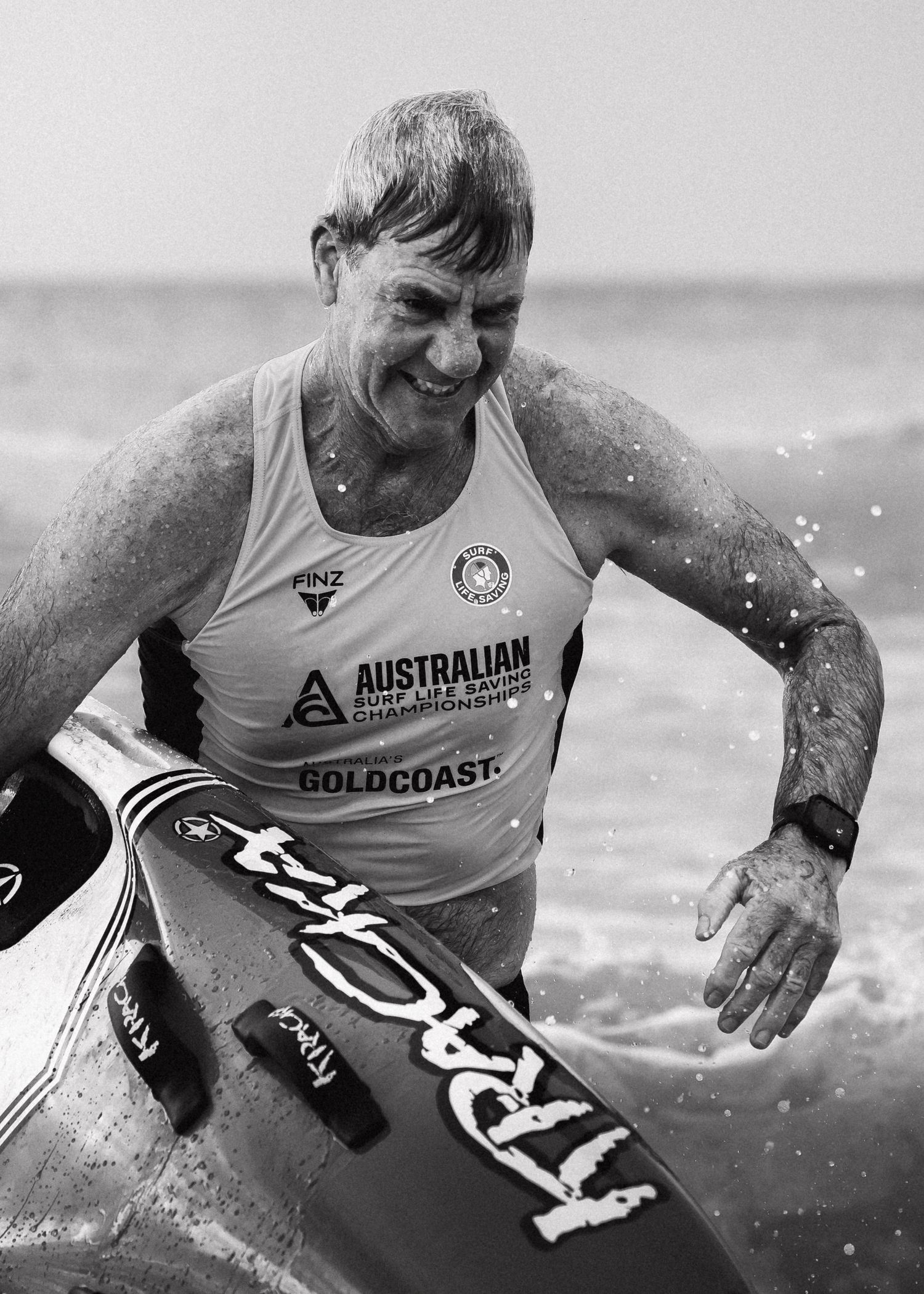 Elderly man smiling, holding a surfboard at Australian Surf Lifesaving Championships on Gold Coast, black and white photo.