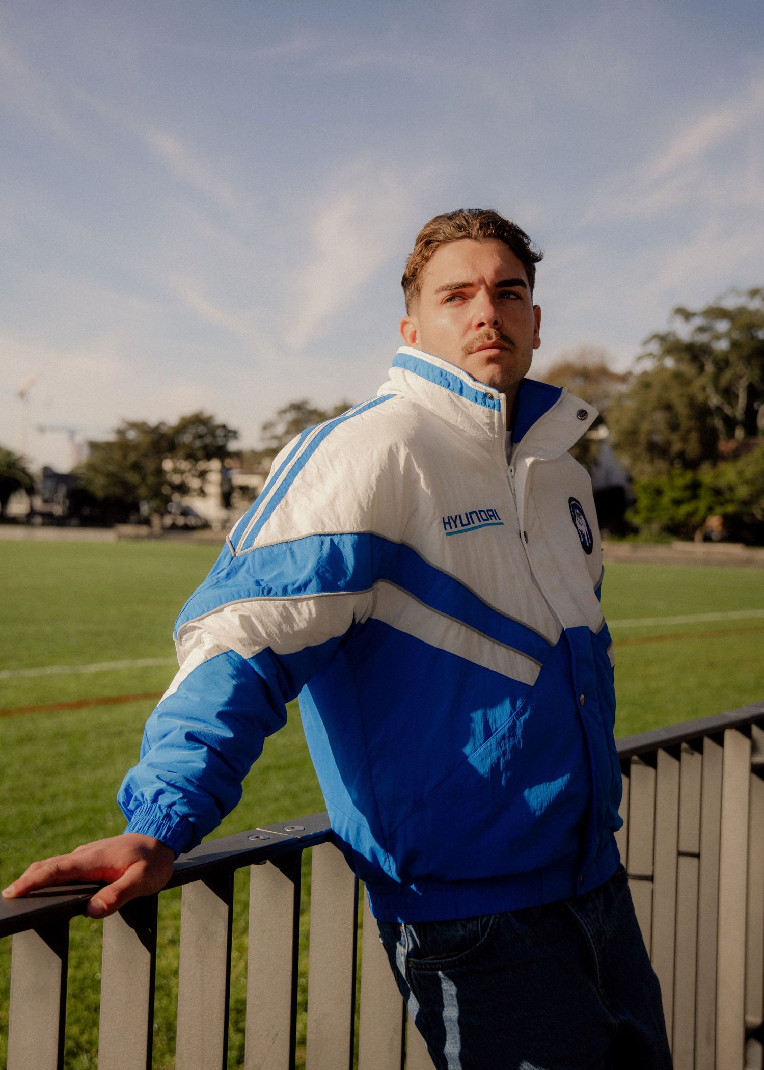 Man in blue and white Hyundai jacket leaning on railing at sports field on sunny day. Sutherland Shire photographer